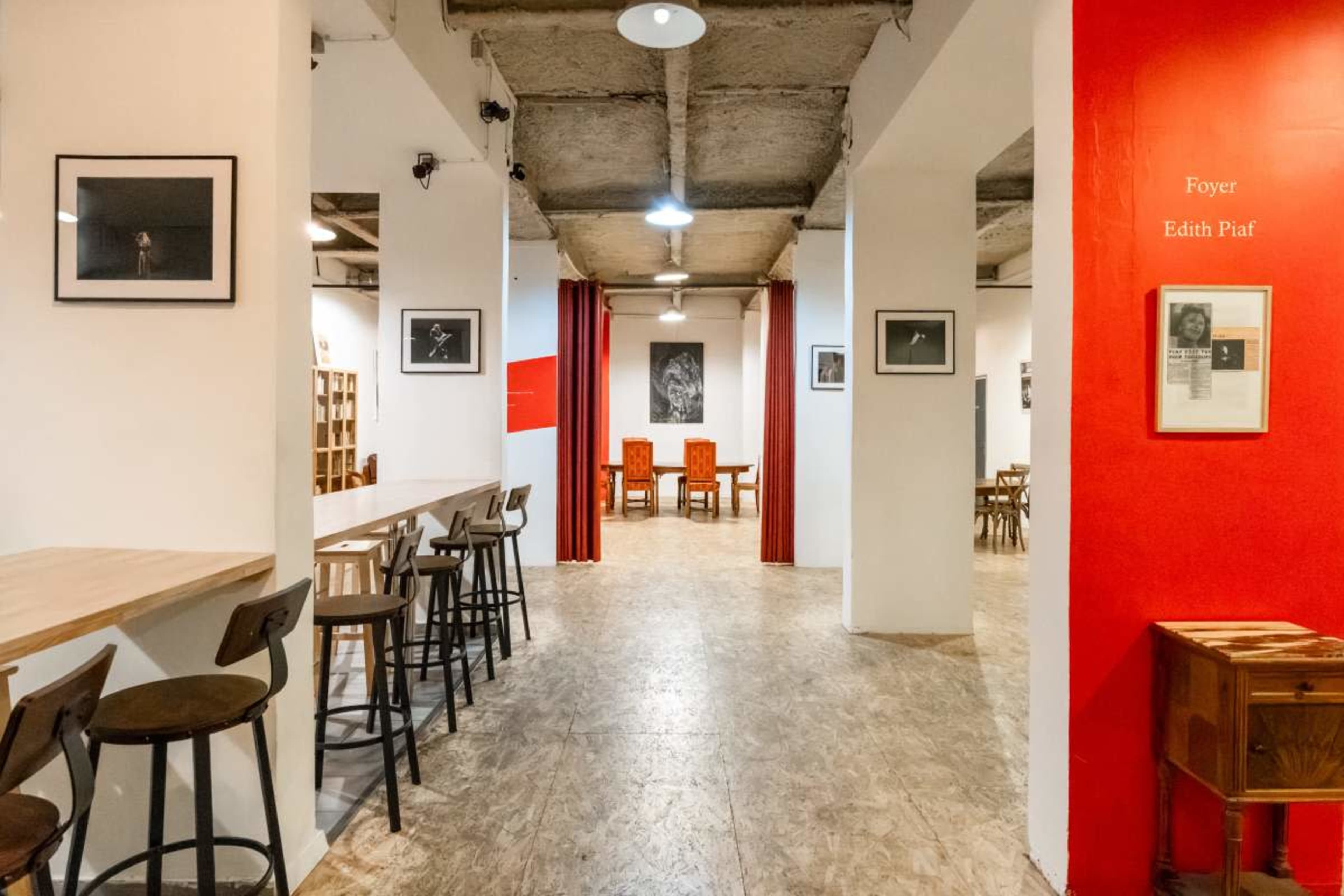 The image shows a spacious art gallery with a wooden bar area on the left and red-accented walls displaying framed photographs, leading to a seating area in the background.