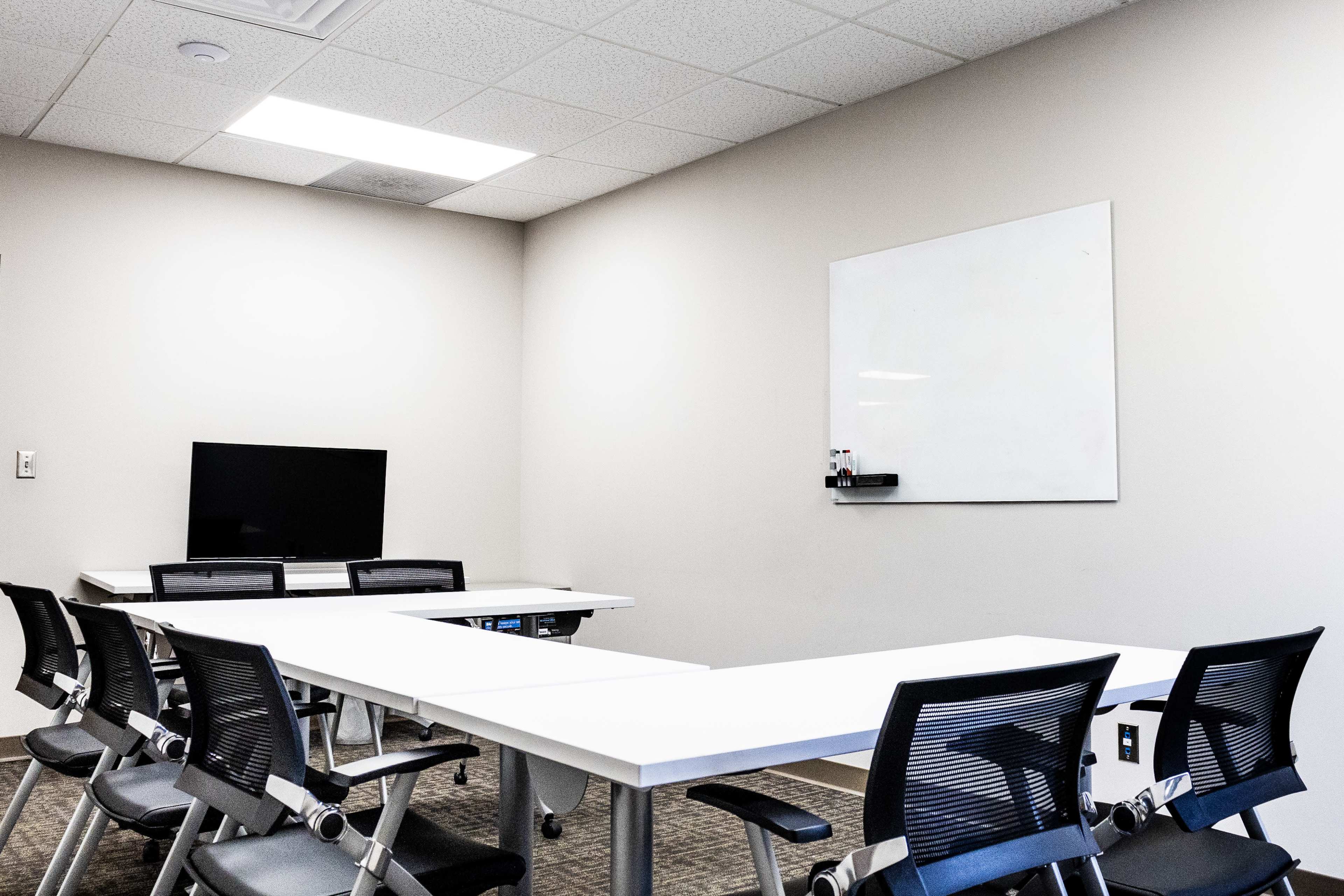 A modern conference room features a rectangular table surrounded by black chairs, a whiteboard on the wall, and a television mounted in one corner.