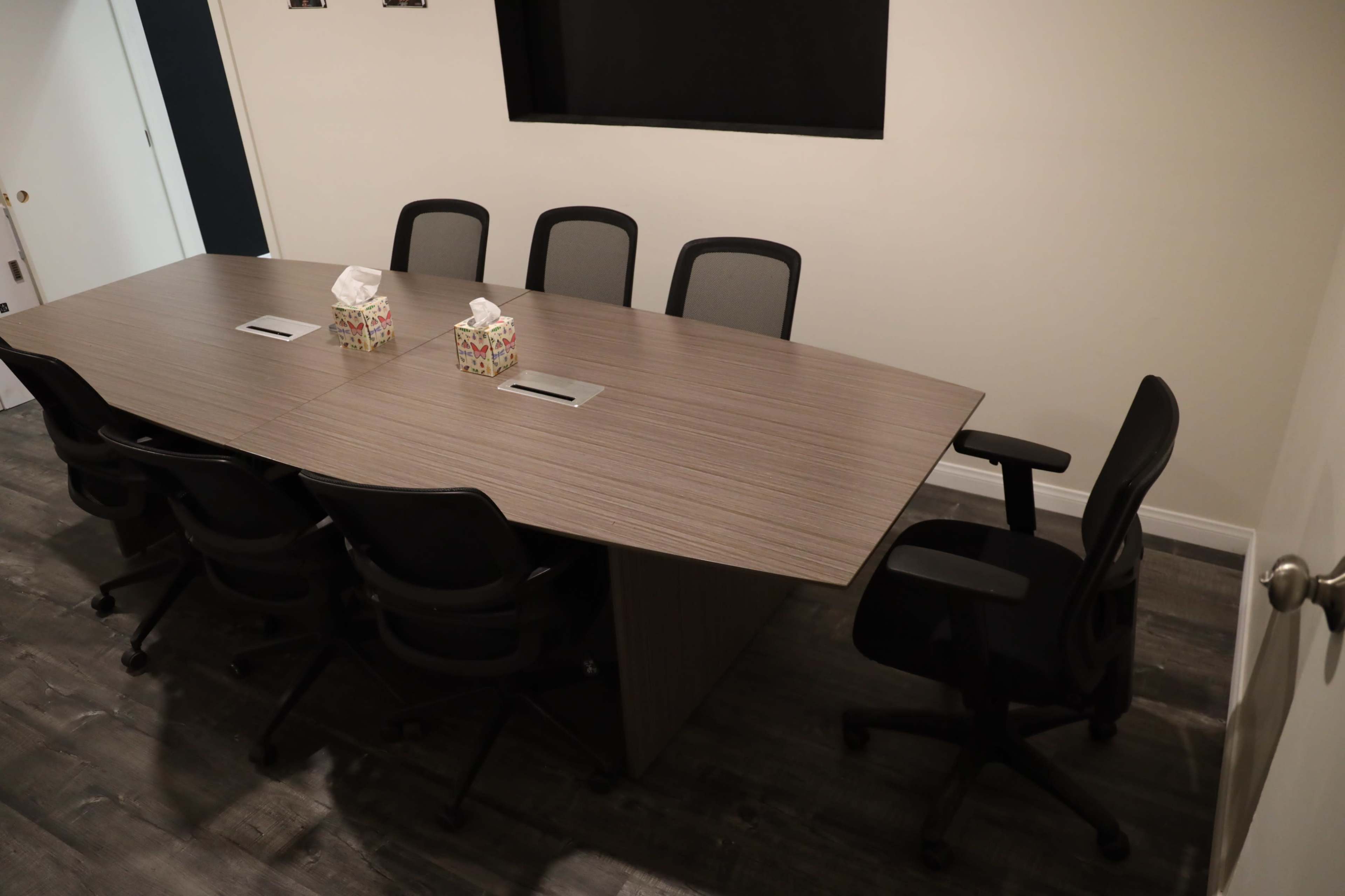 A conference room features a large, rectangular table surrounded by seven black office chairs, with two tissue boxes placed on the table's surface.