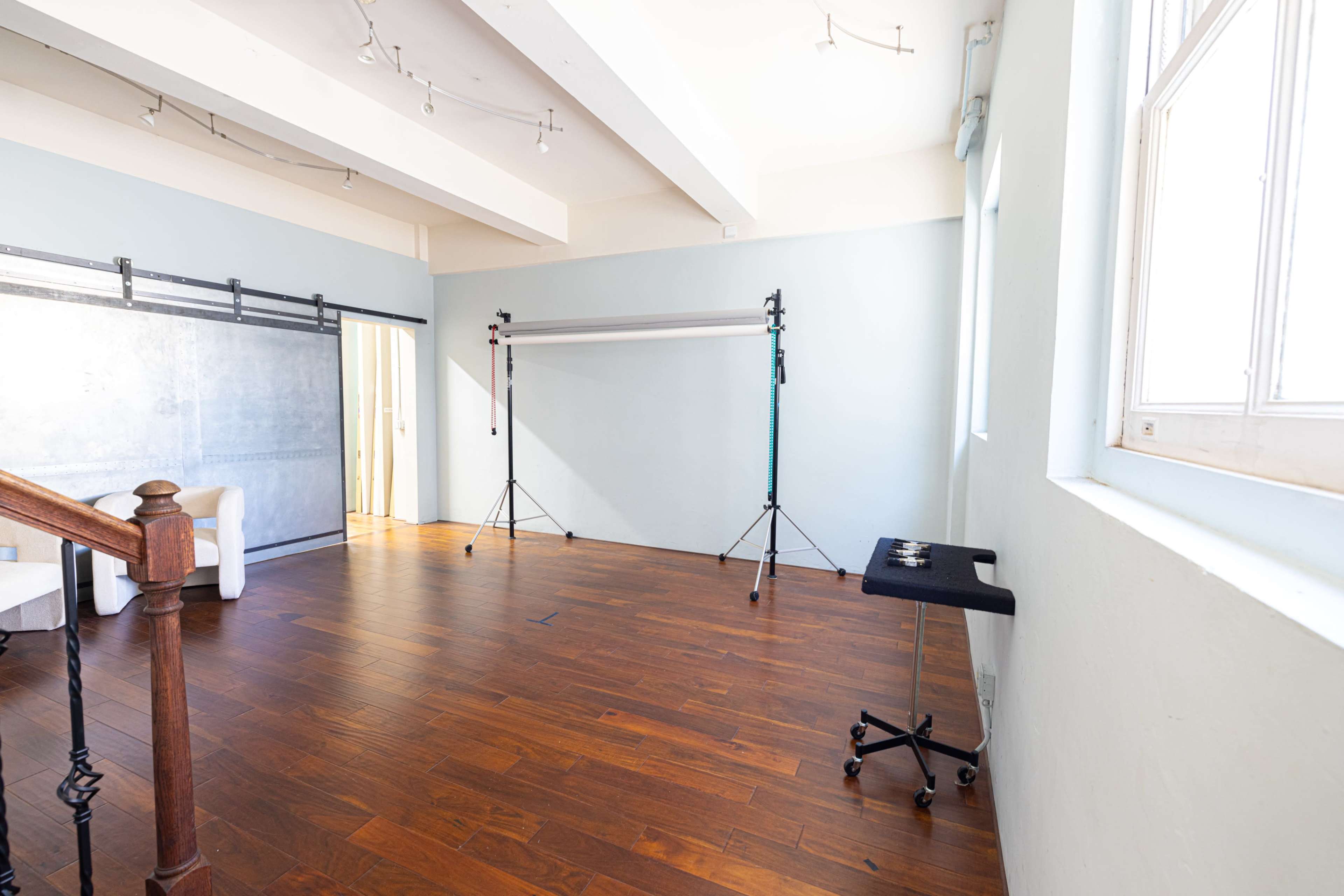 The image shows a spacious, well-lit room with wooden flooring, featuring a simple setup of a backdrop stand and two light stands.