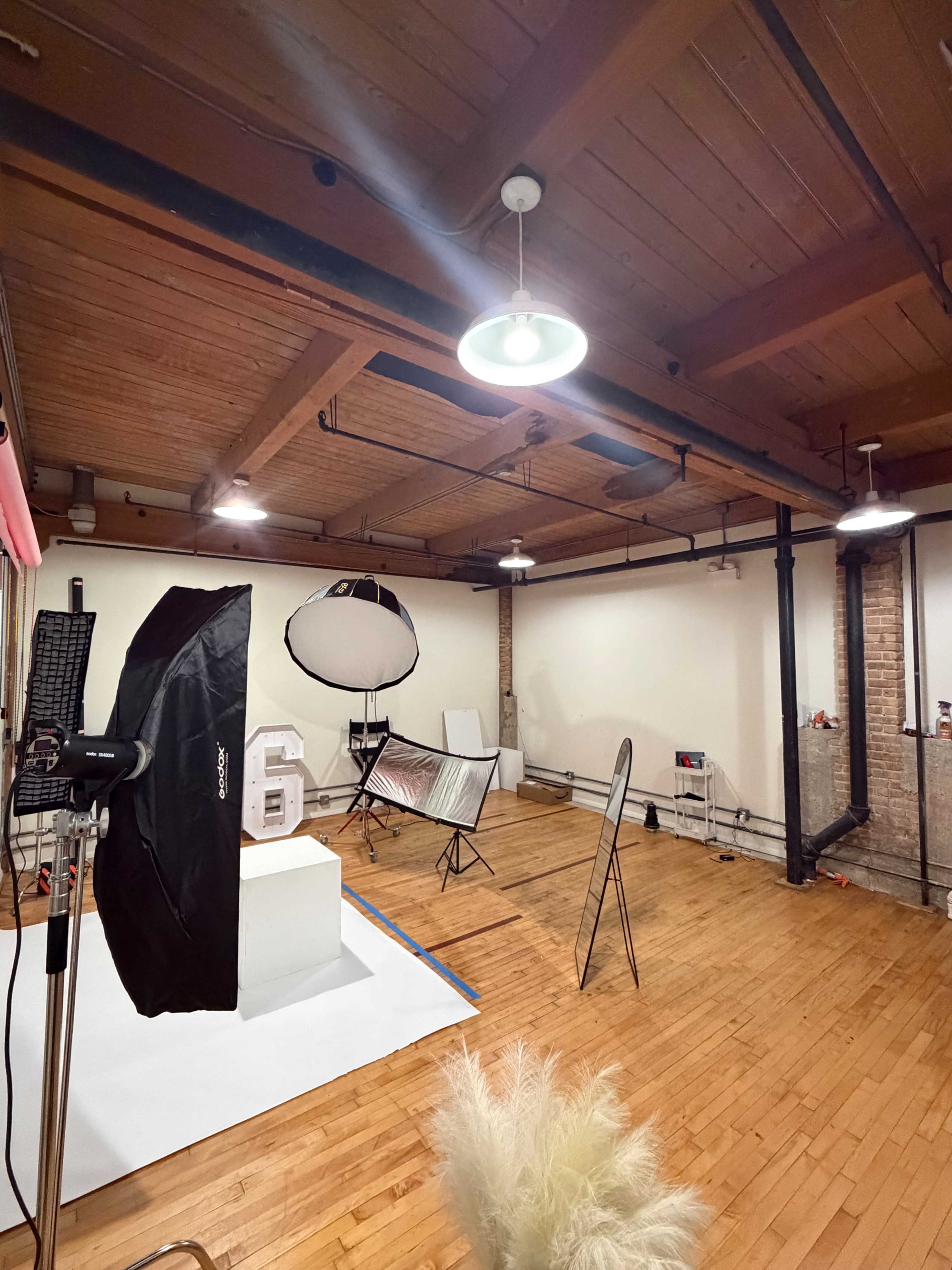 The image shows a spacious photography studio with wooden floors, studio lighting equipment, and various backdrops set up for a photoshoot.