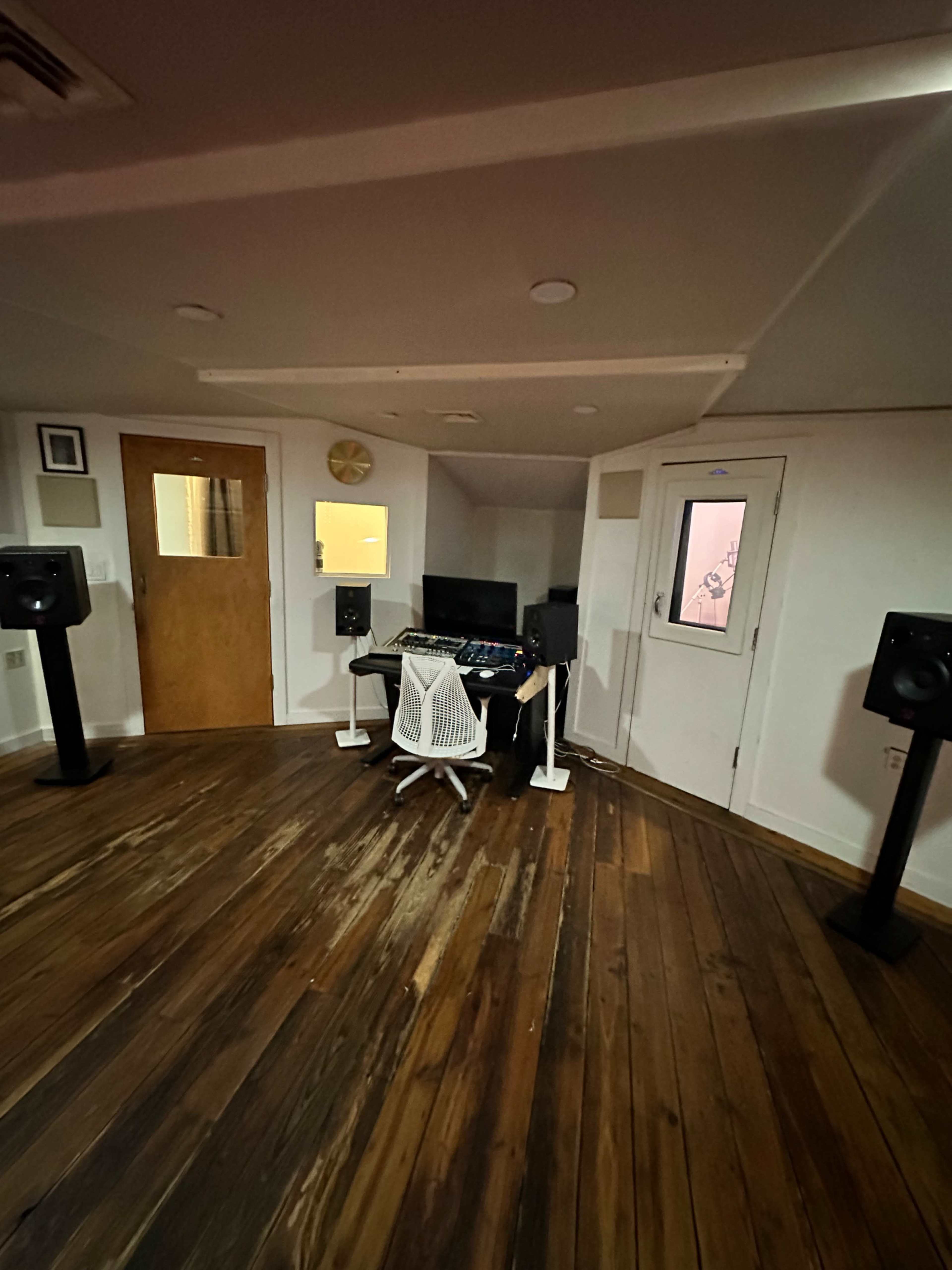 A home studio features a desk with a computer and audio equipment, surrounded by tall speakers on either side, with wooden flooring and a door in the background.