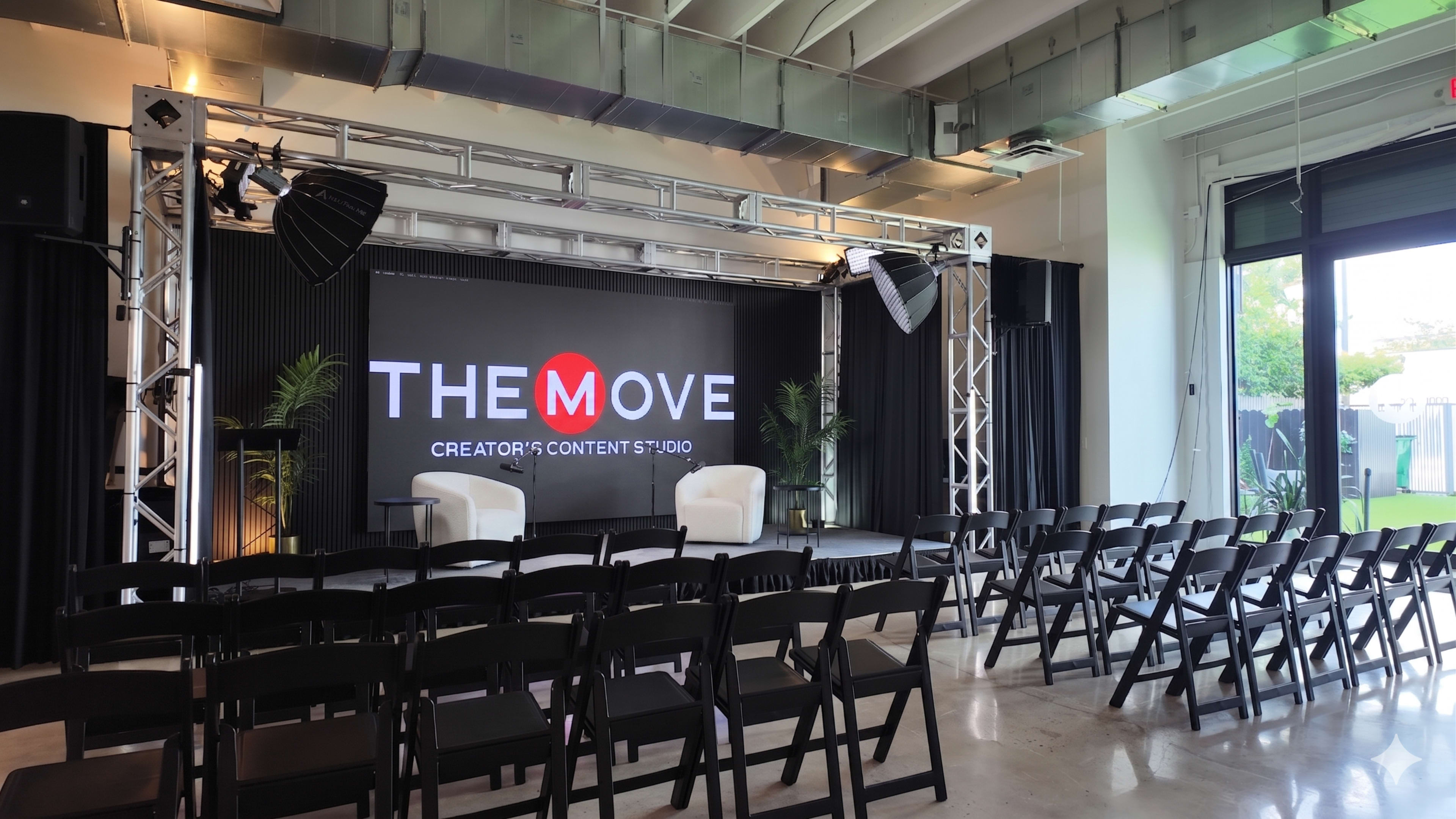 The image shows a modern studio setup with two white chairs on a stage, flanked by black curtains and a large LED screen displaying "THE MOVE CREATOR'S CONTENT STUDIO." Rows of black folding chairs are arranged in front of the stage.