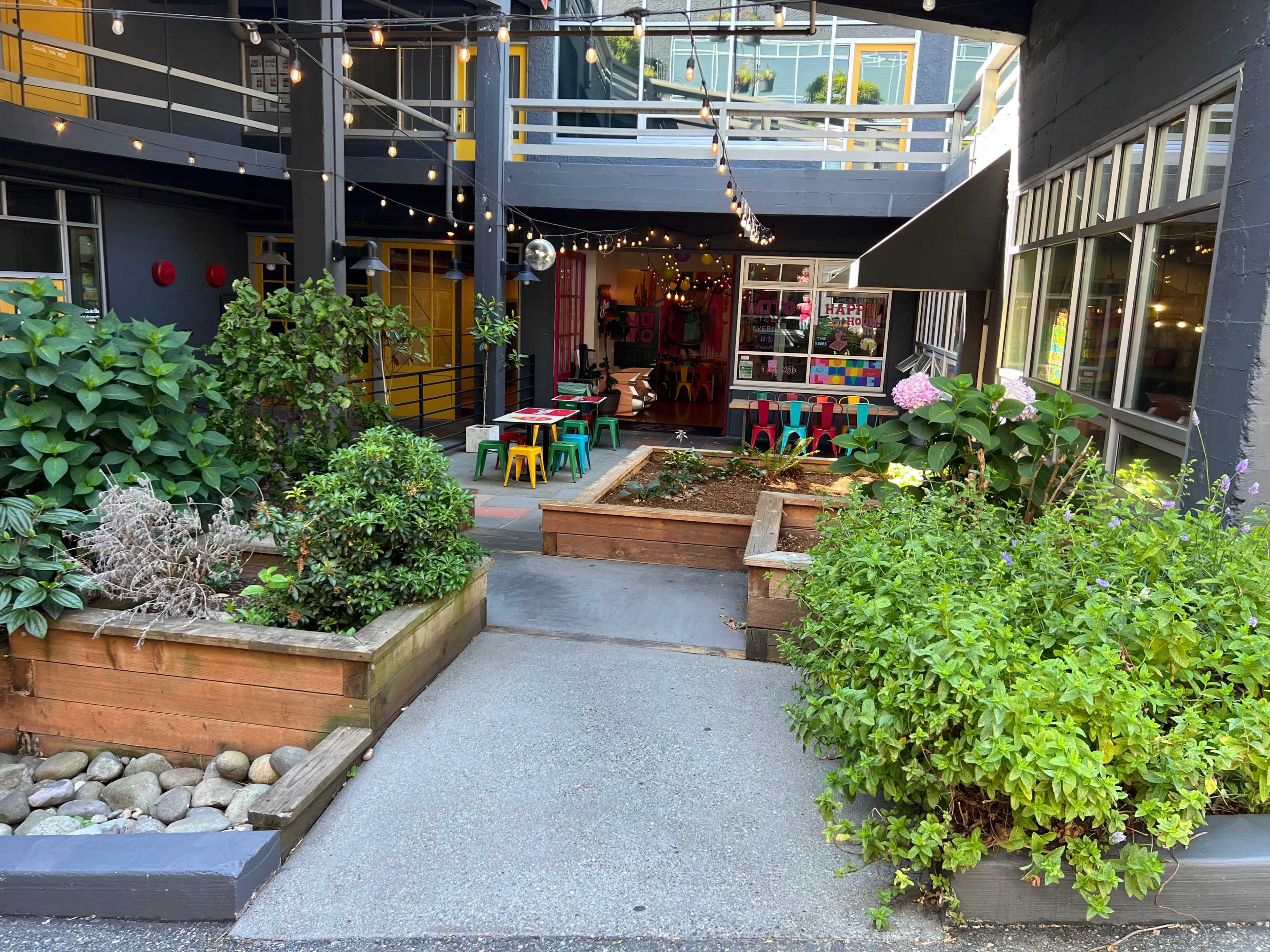 The image shows a landscaped courtyard area featuring raised garden beds and a path leading to a colorful outdoor seating area with string lights overhead.