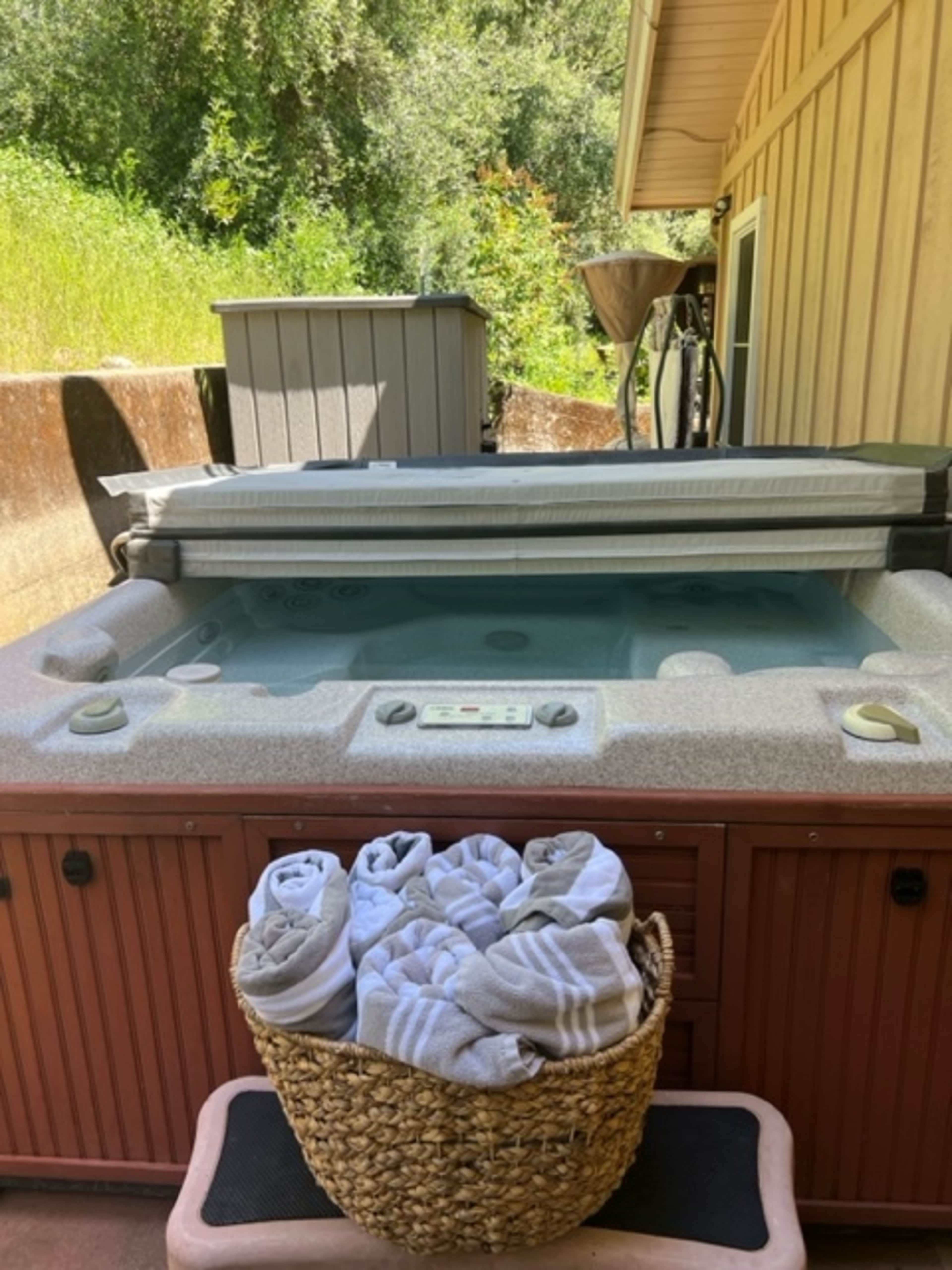 A hot tub with a cover and a basket of towels sits beside a wooden deck and a shed, surrounded by greenery.
