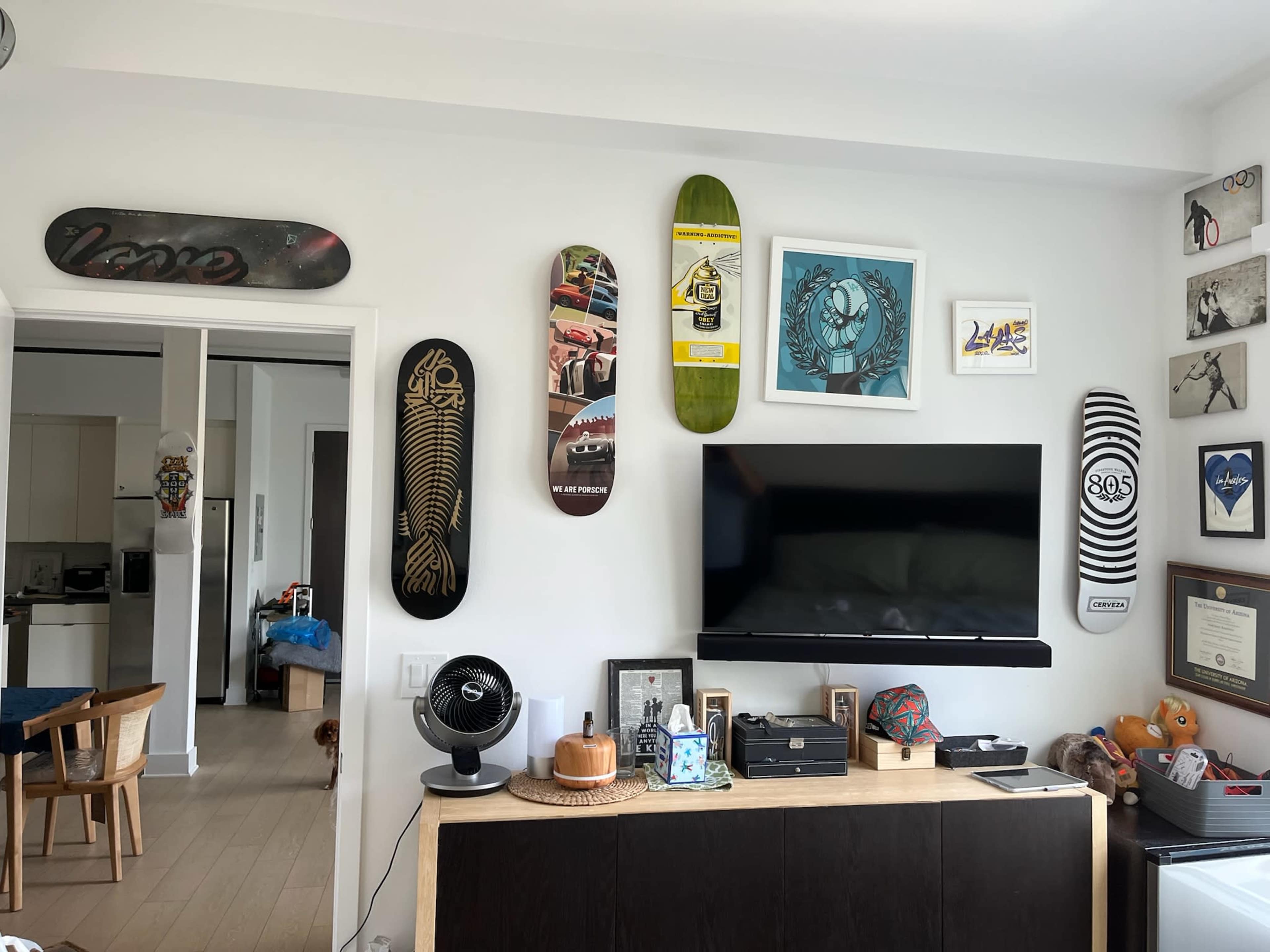 The image shows a living room with a television mounted on a wall, surrounded by decorative skateboards and framed art, while a dining area is visible in the background.