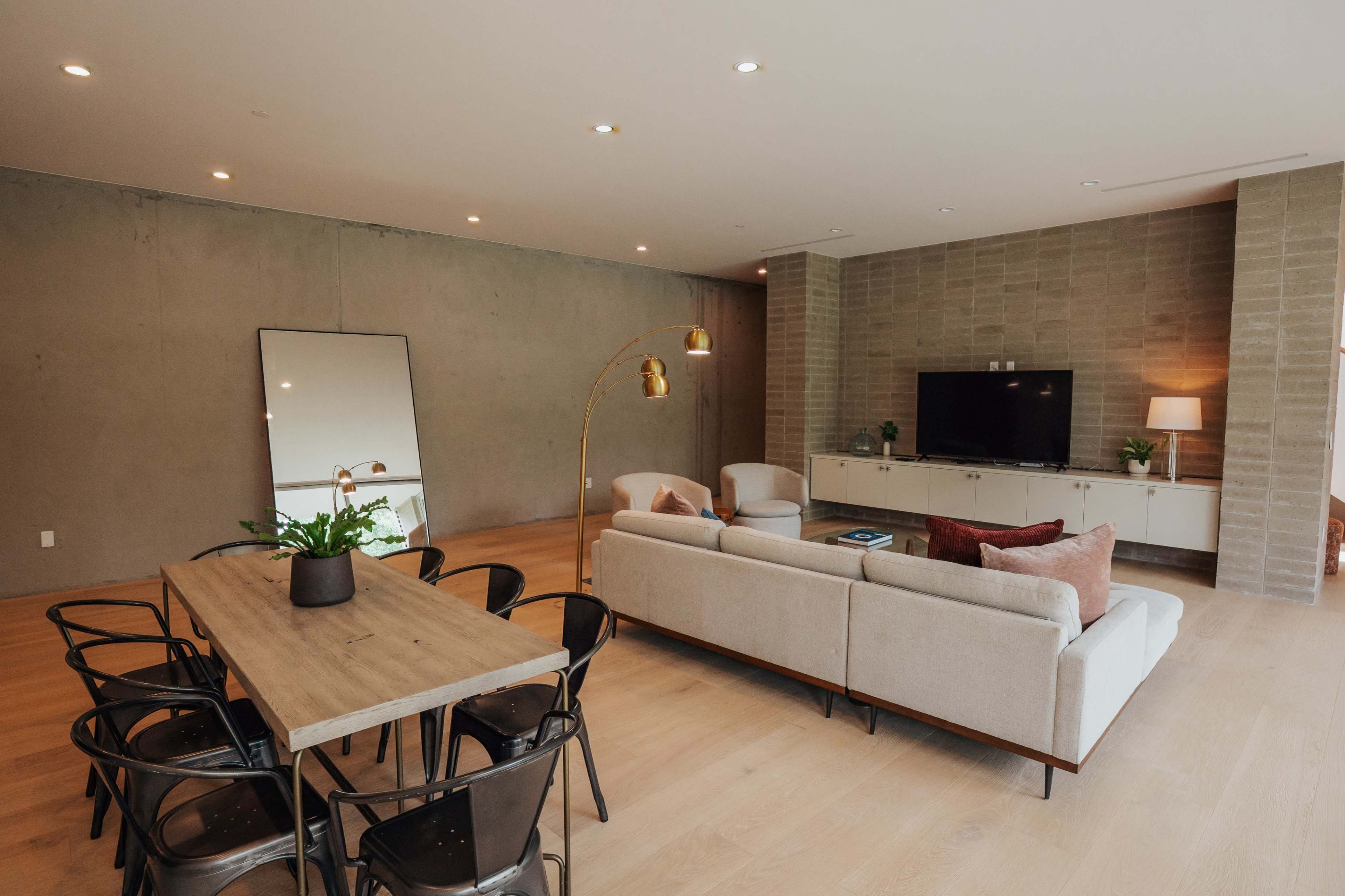 The image shows a modern living space featuring a dining table with black chairs, a sectional sofa, a television mounted on a wall, and a large mirror against a concrete backdrop.