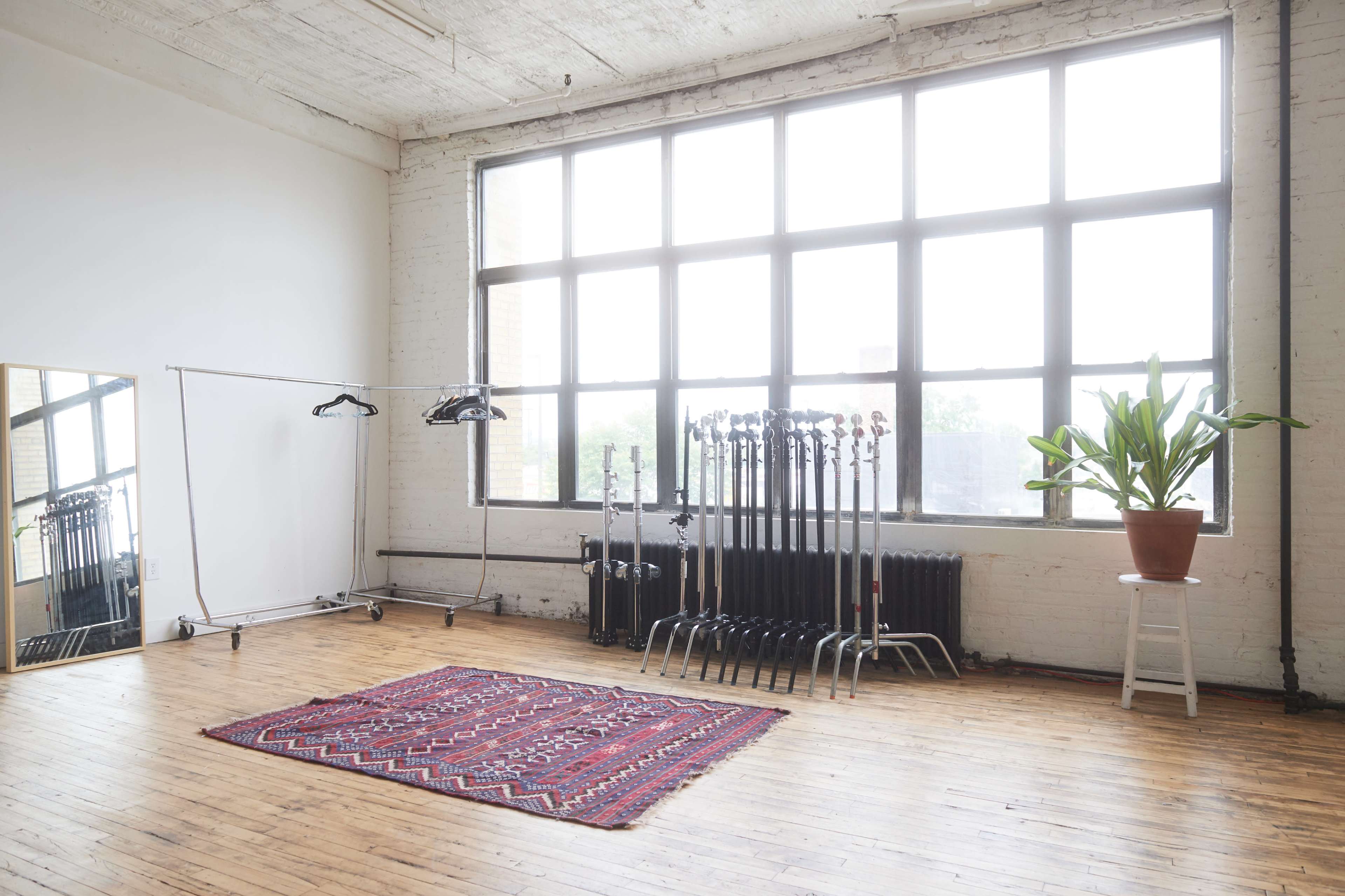 A spacious, well-lit room featuring large windows, a patterned rug on the wooden floor, a mirror on wheels, and several clothing racks with hangers.