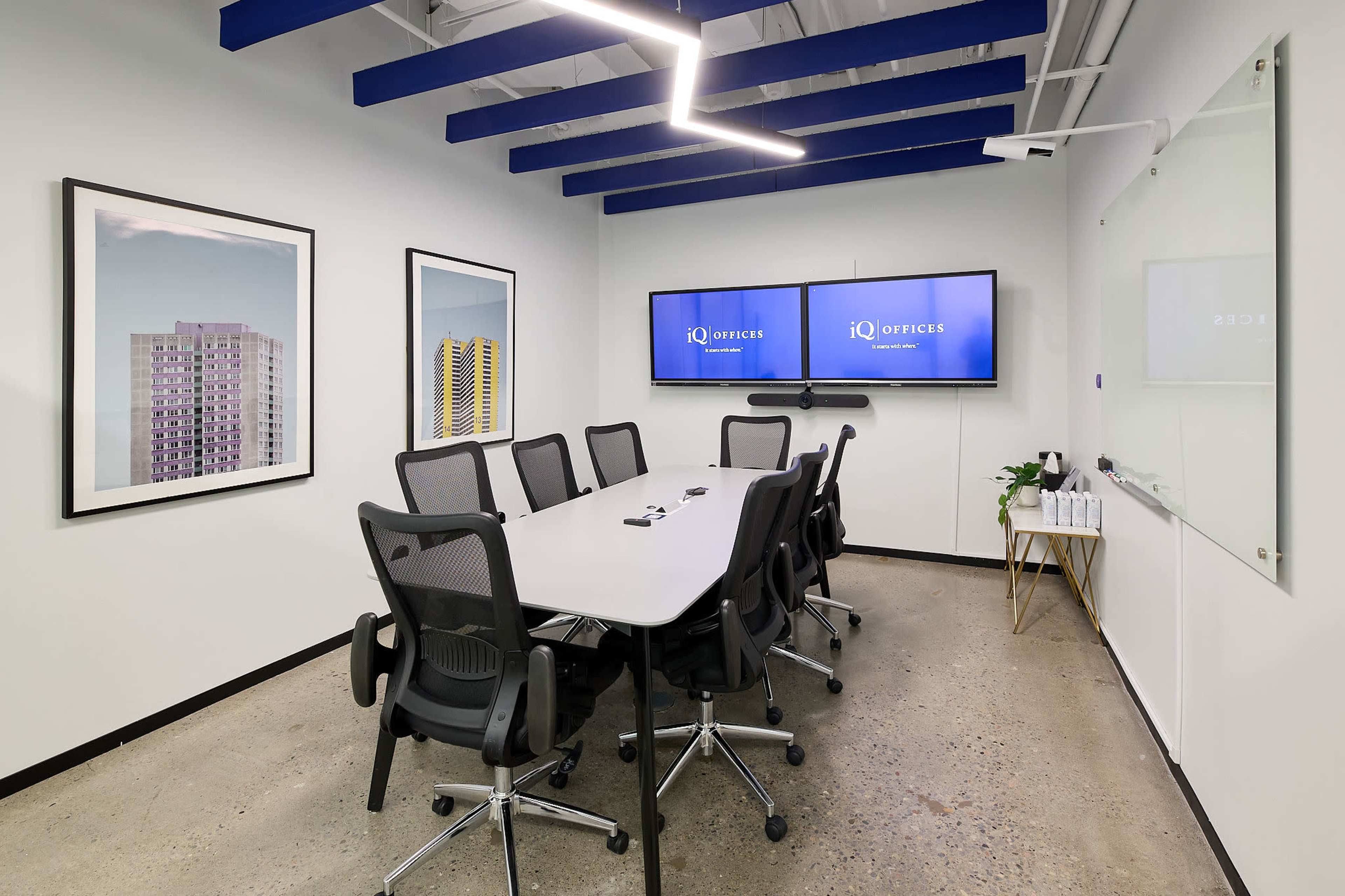 The image shows a modern conference room with a long table, multiple ergonomic chairs, two large screens displaying the logo "IQ Offices," and a whiteboard on one wall.