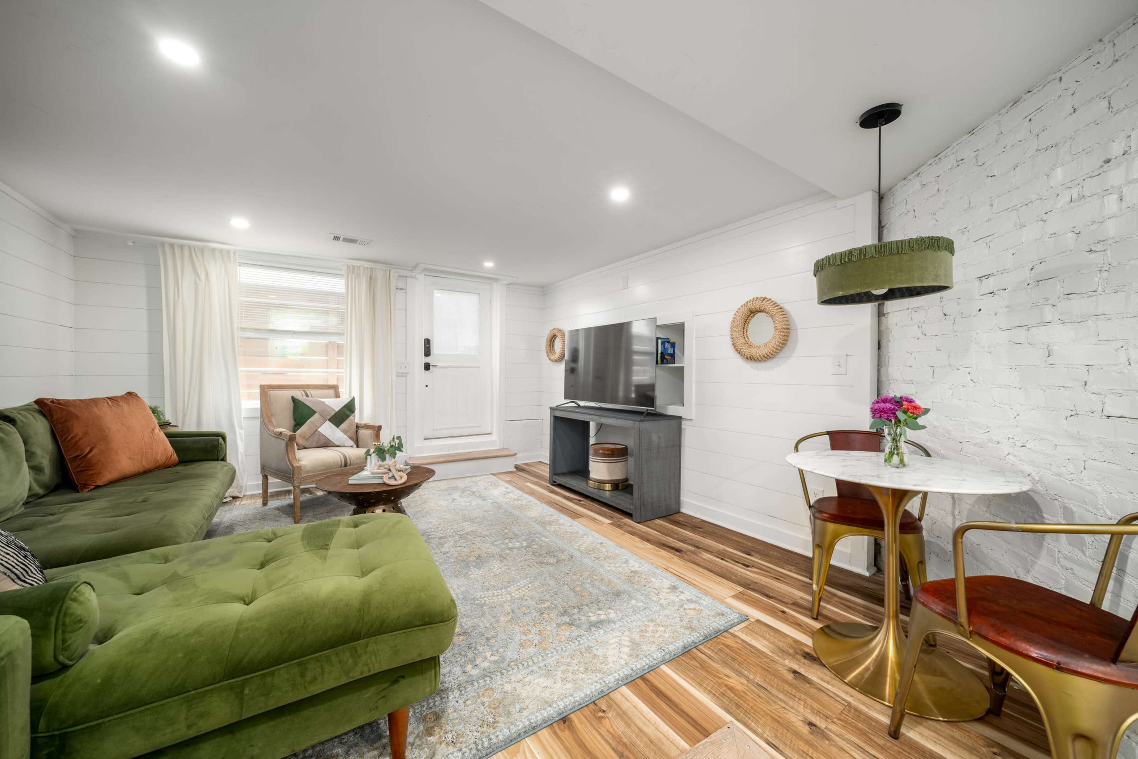 The image shows a cozy living room featuring a green sofa, a small round dining table with two chairs, and a television mounted on the wall.