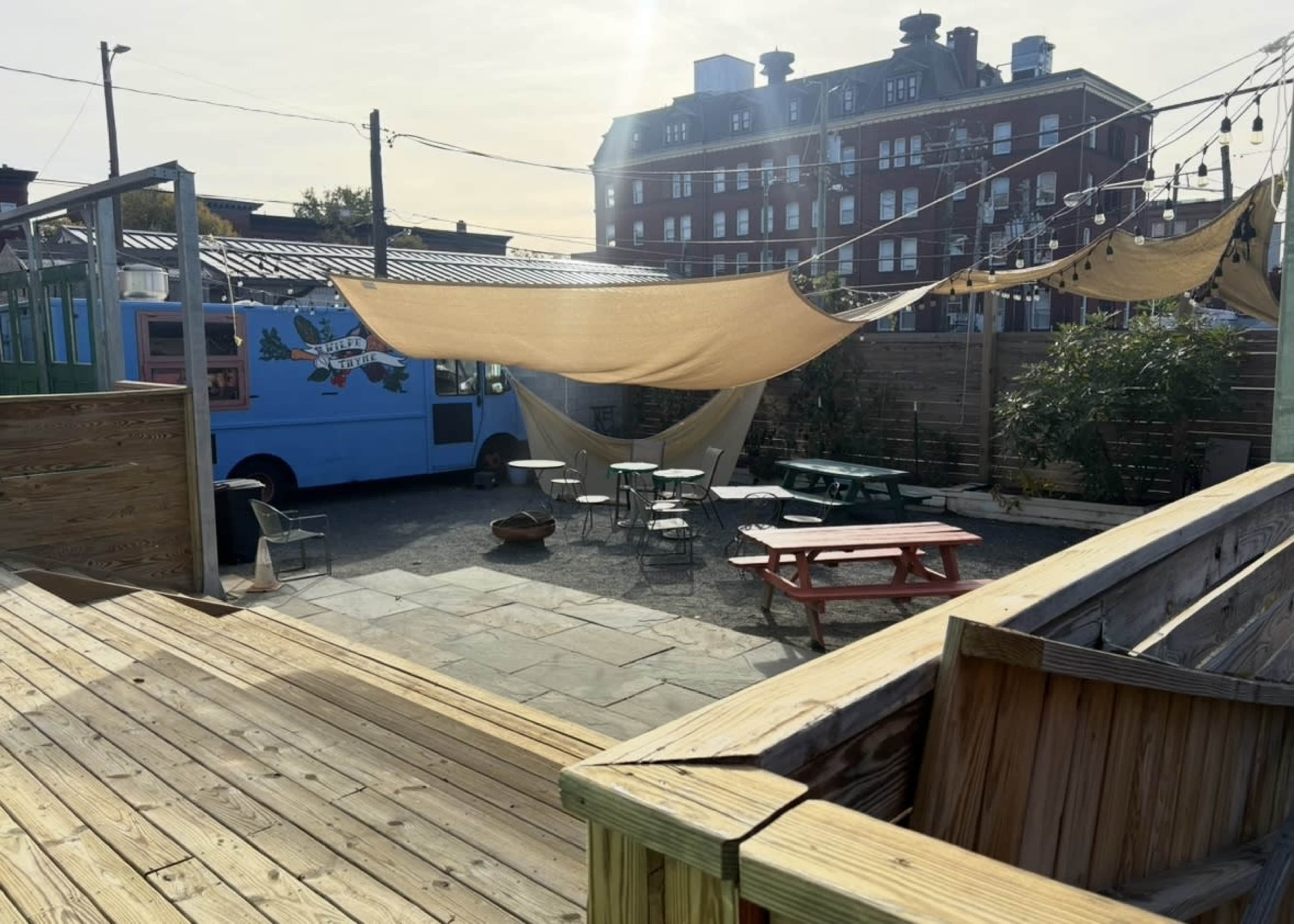 The image shows an outdoor seating area with picnic tables, a colorful food truck, and shade sails around a gravel lot, set against a backdrop of a brick building.