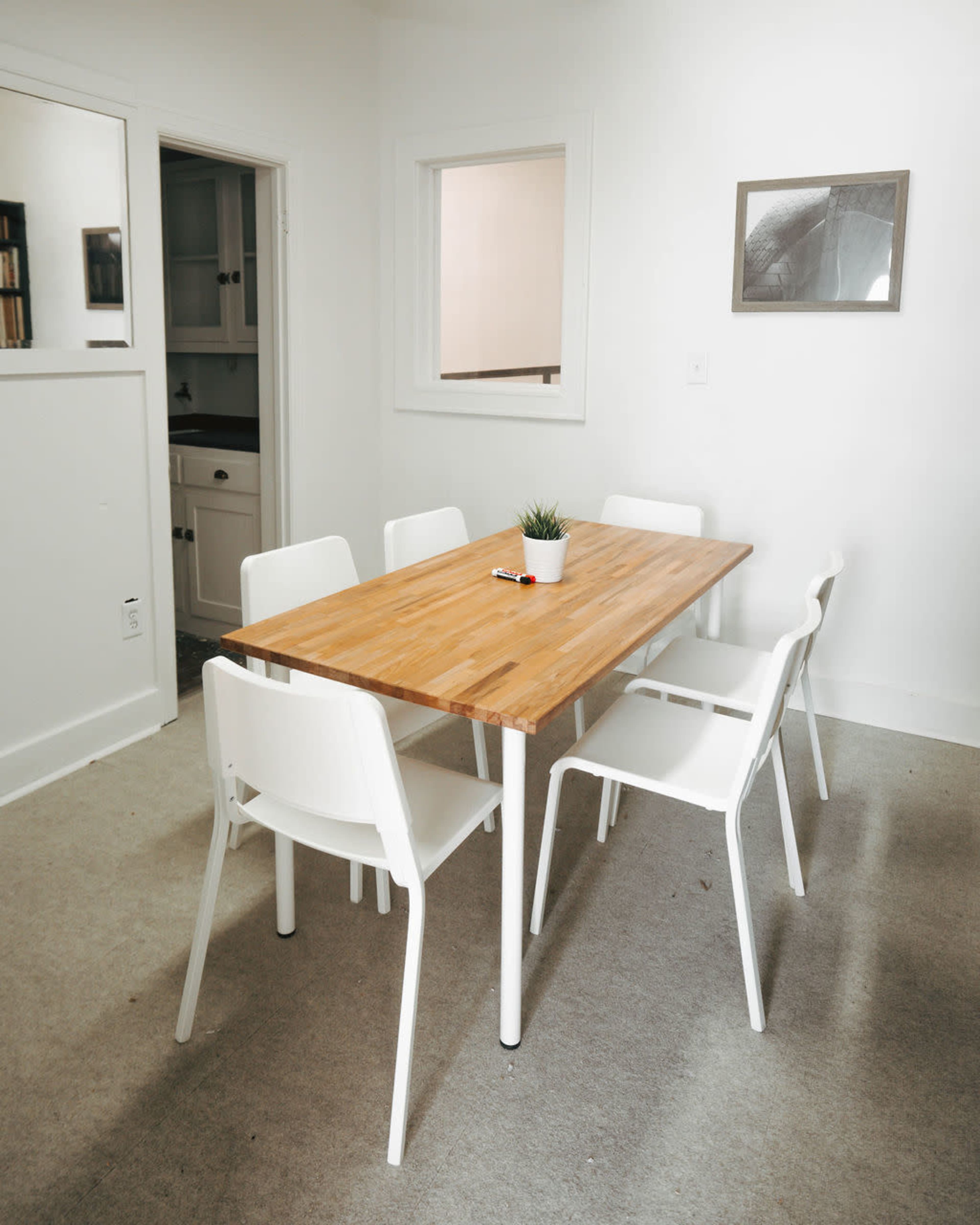 A wooden dining table surrounded by six white chairs is set in a bright, minimalistic room with light-colored walls and a small plant centerpiece.