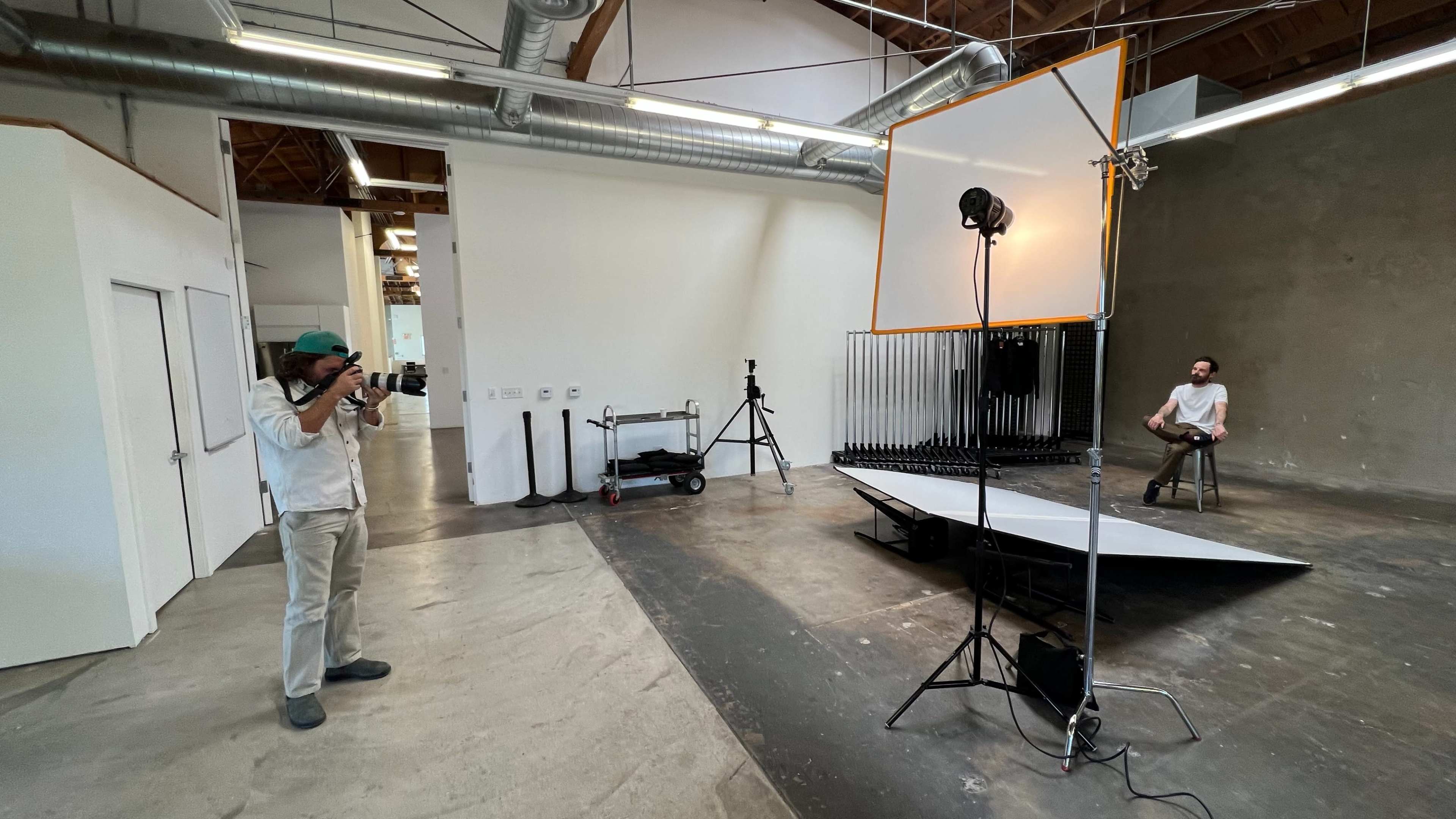 A photographer is setting up a shoot in a spacious studio with a model sitting on a stool in front of a light setup.