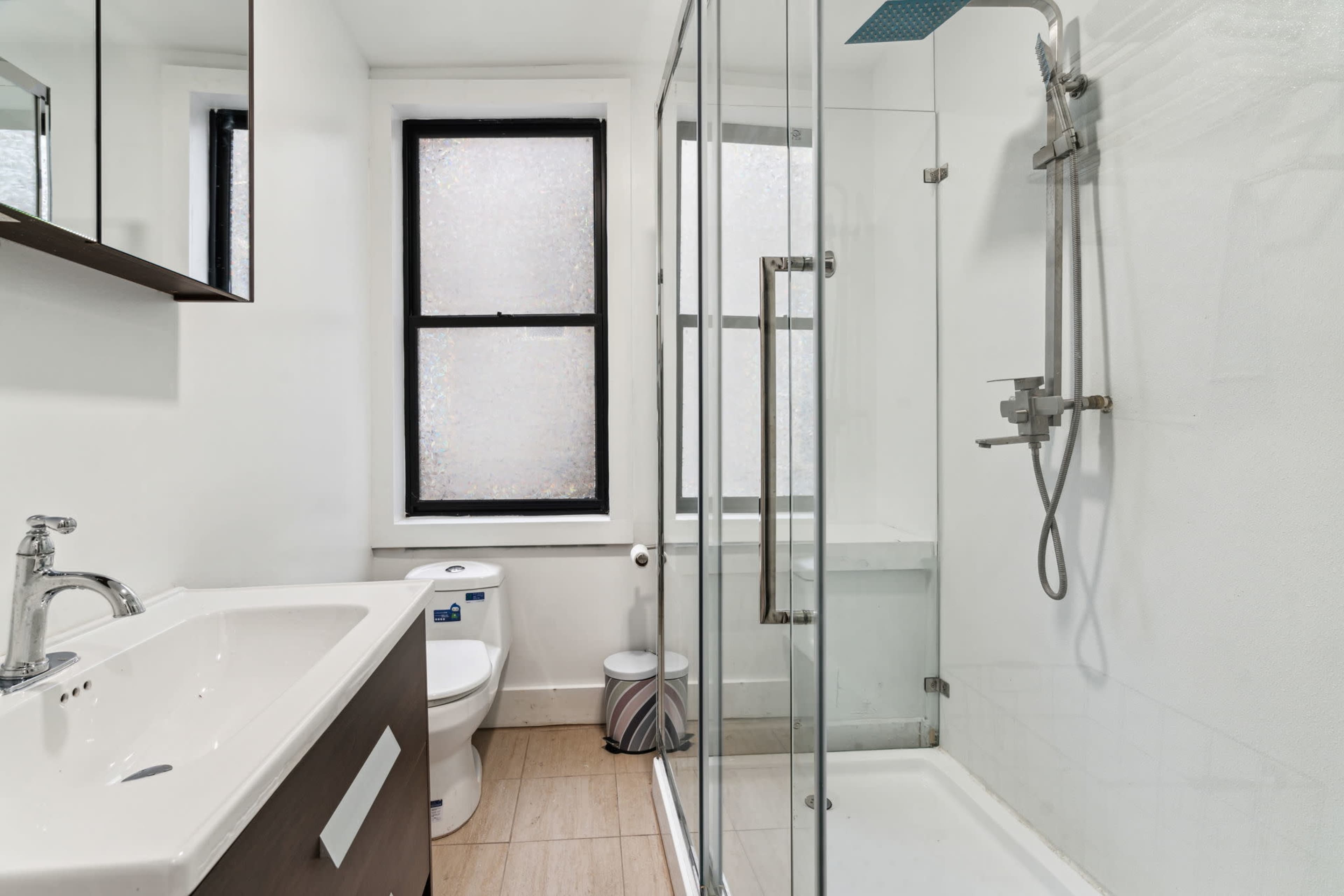 The image shows a modern bathroom with a glass shower enclosure, a toilet, a sink with a dark wood vanity, and large windows.