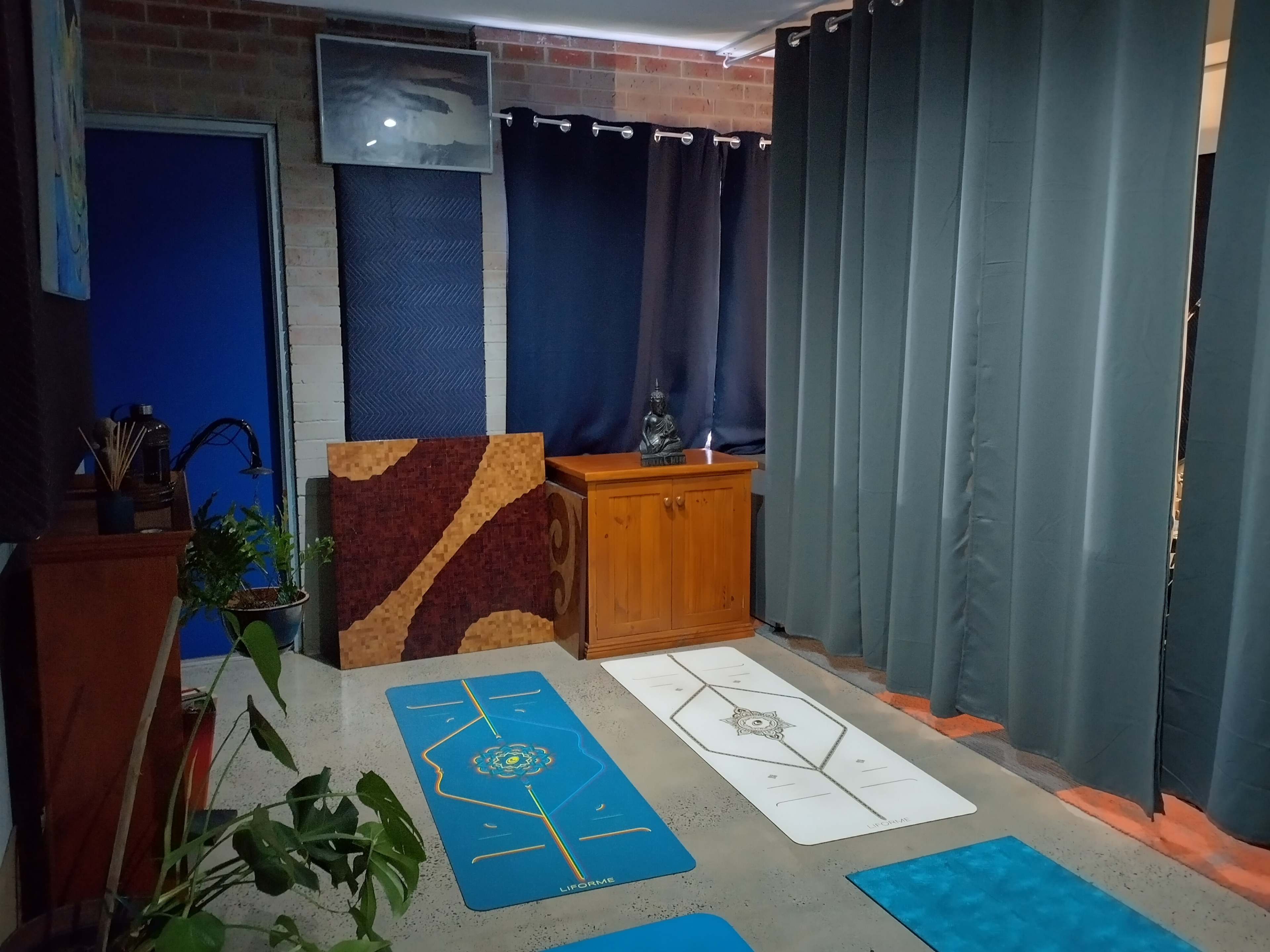 A yoga studio with colorful mats arranged on the floor, a wooden cabinet, and a statue, all set against a backdrop of dark curtains and a brick wall.