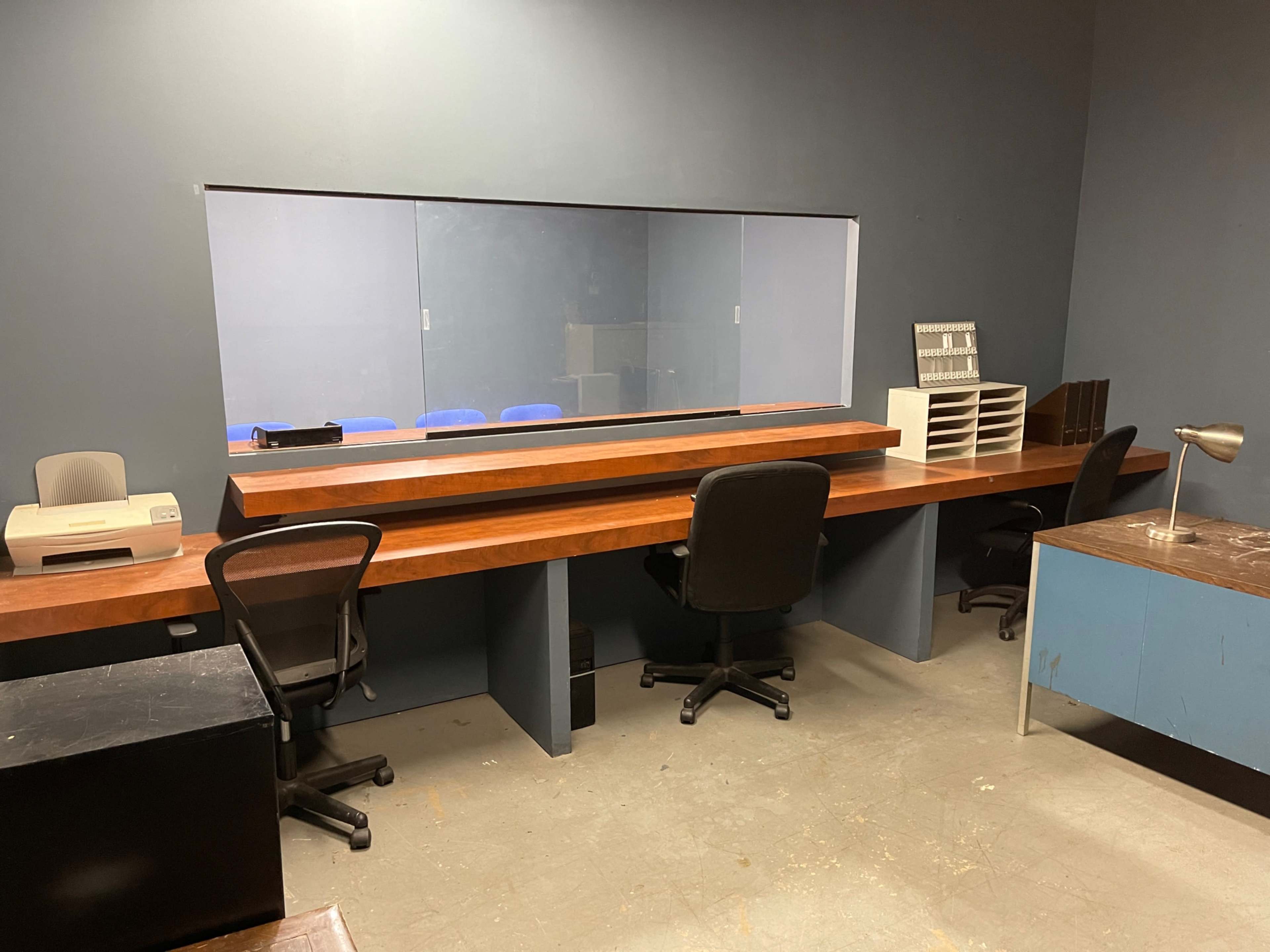 The image shows an office space with two desks, a printer, and a large window facing another room.