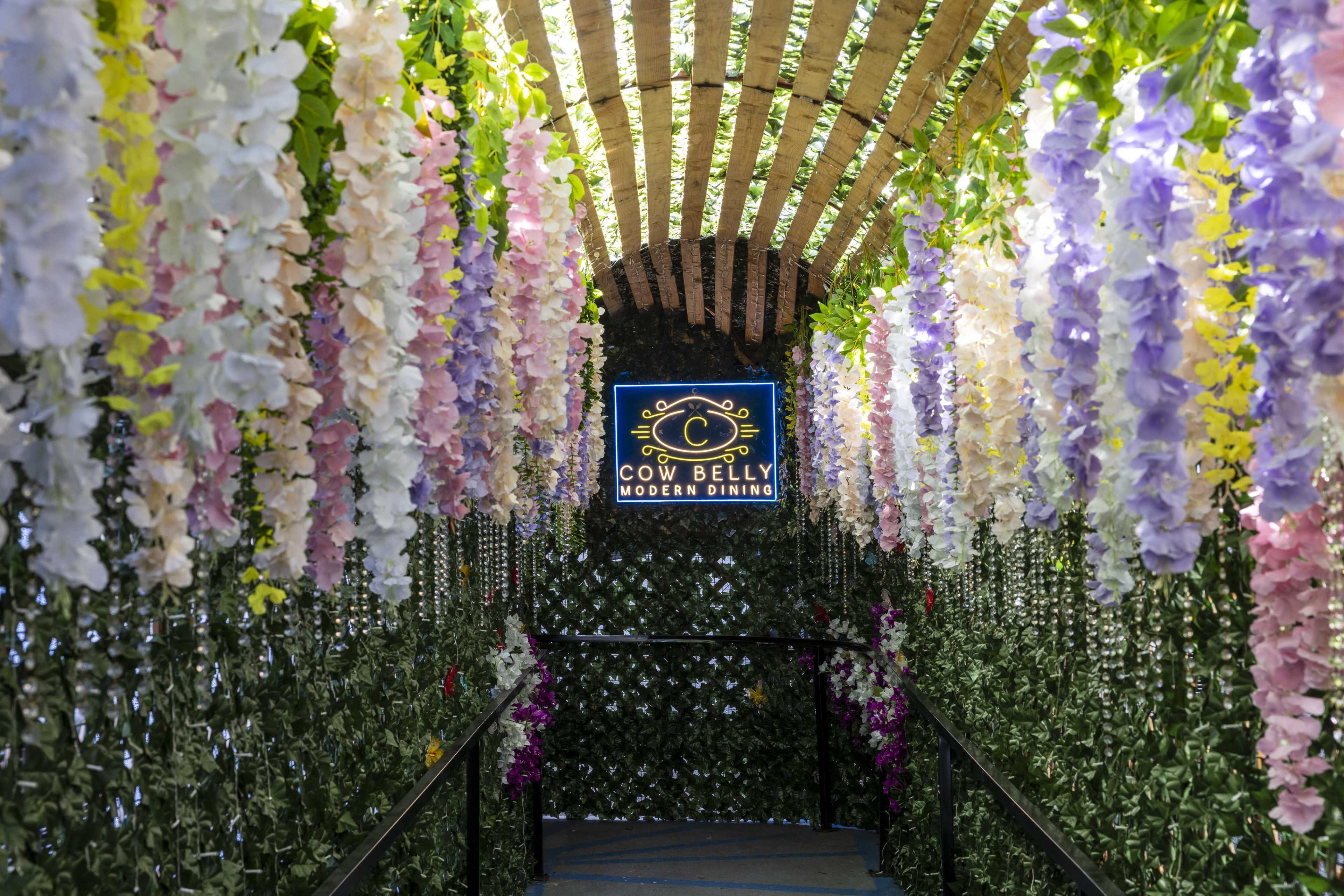 The image depicts a floral archway adorned with colorful hanging flowers leading to a sign that reads "Cow Belly Modern Dining."