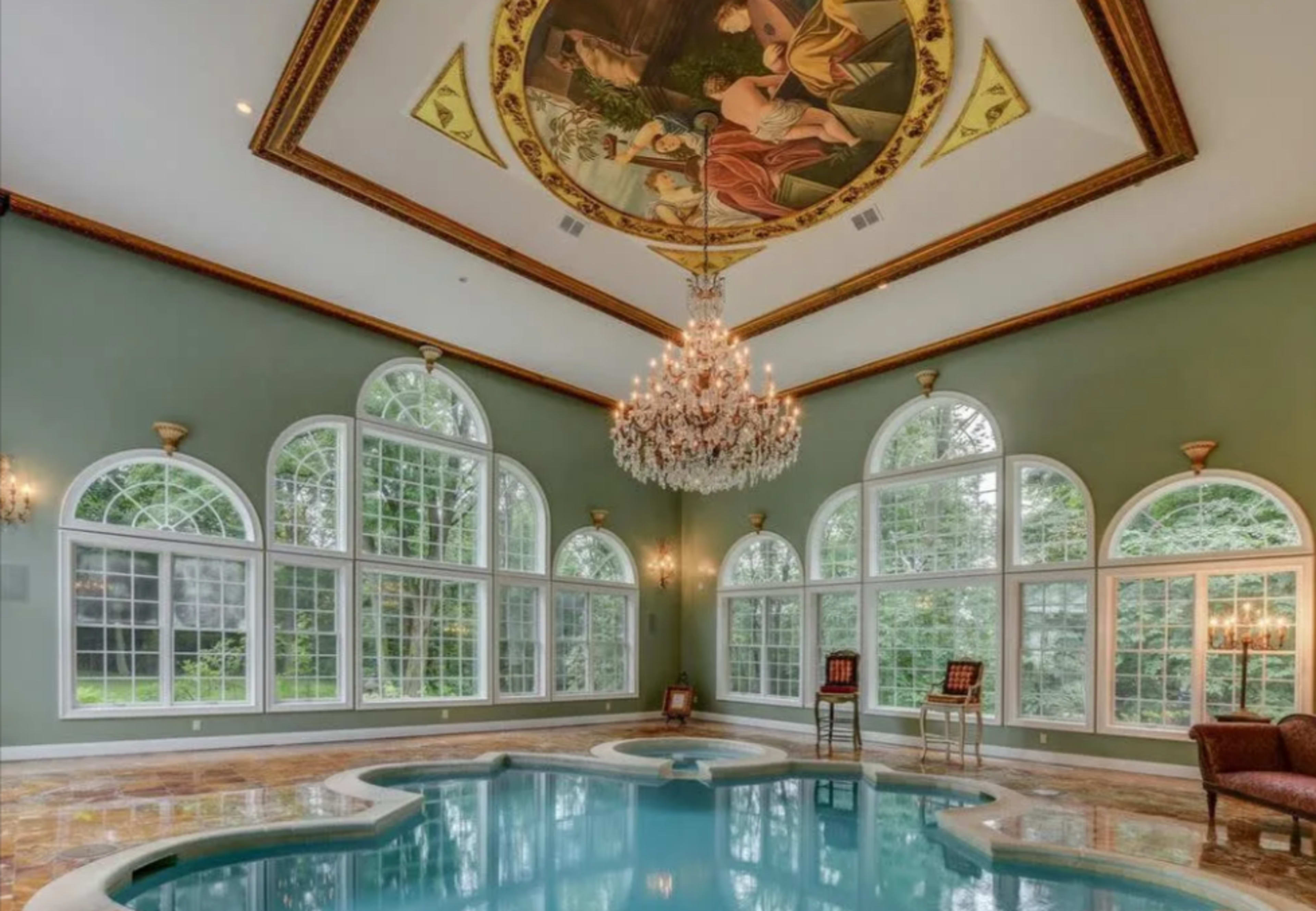A luxurious indoor swimming pool area with large arched windows, a chandelier, and a painted ceiling.