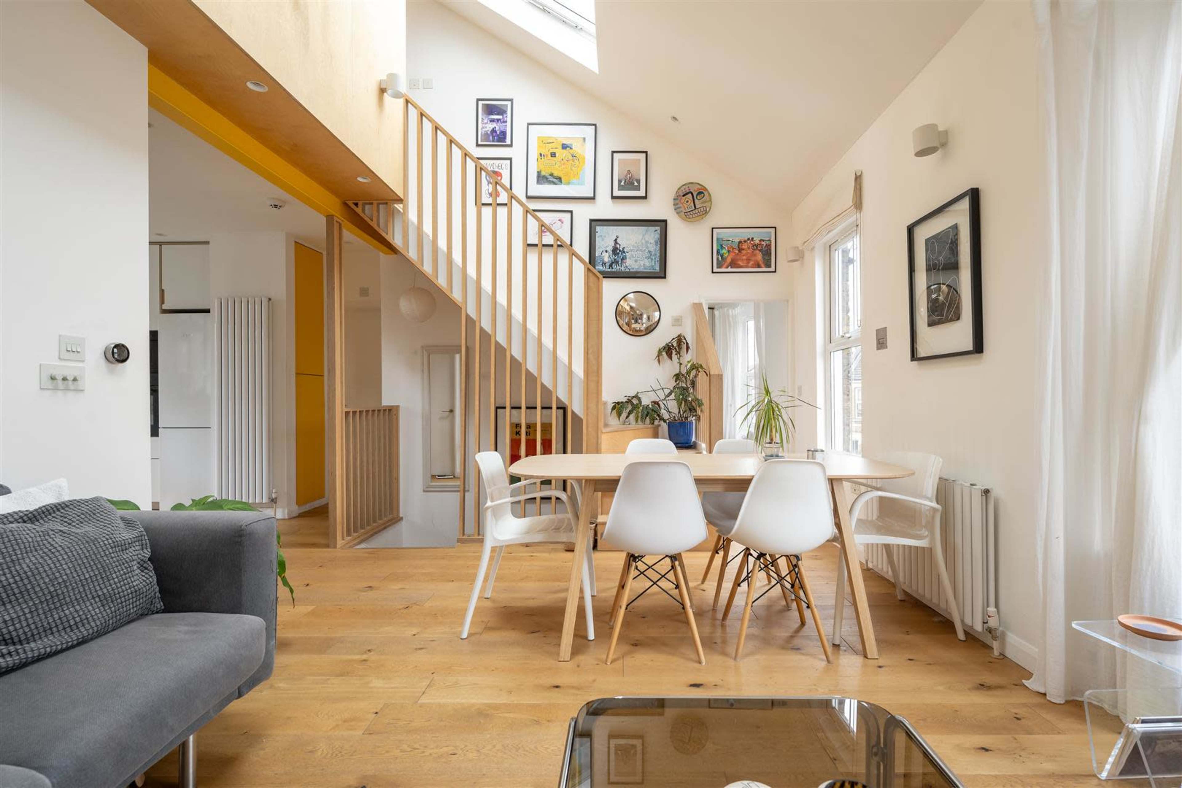 The image shows a modern interior space featuring a dining table surrounded by white chairs, with a staircase in the background and artwork hanging on the walls.