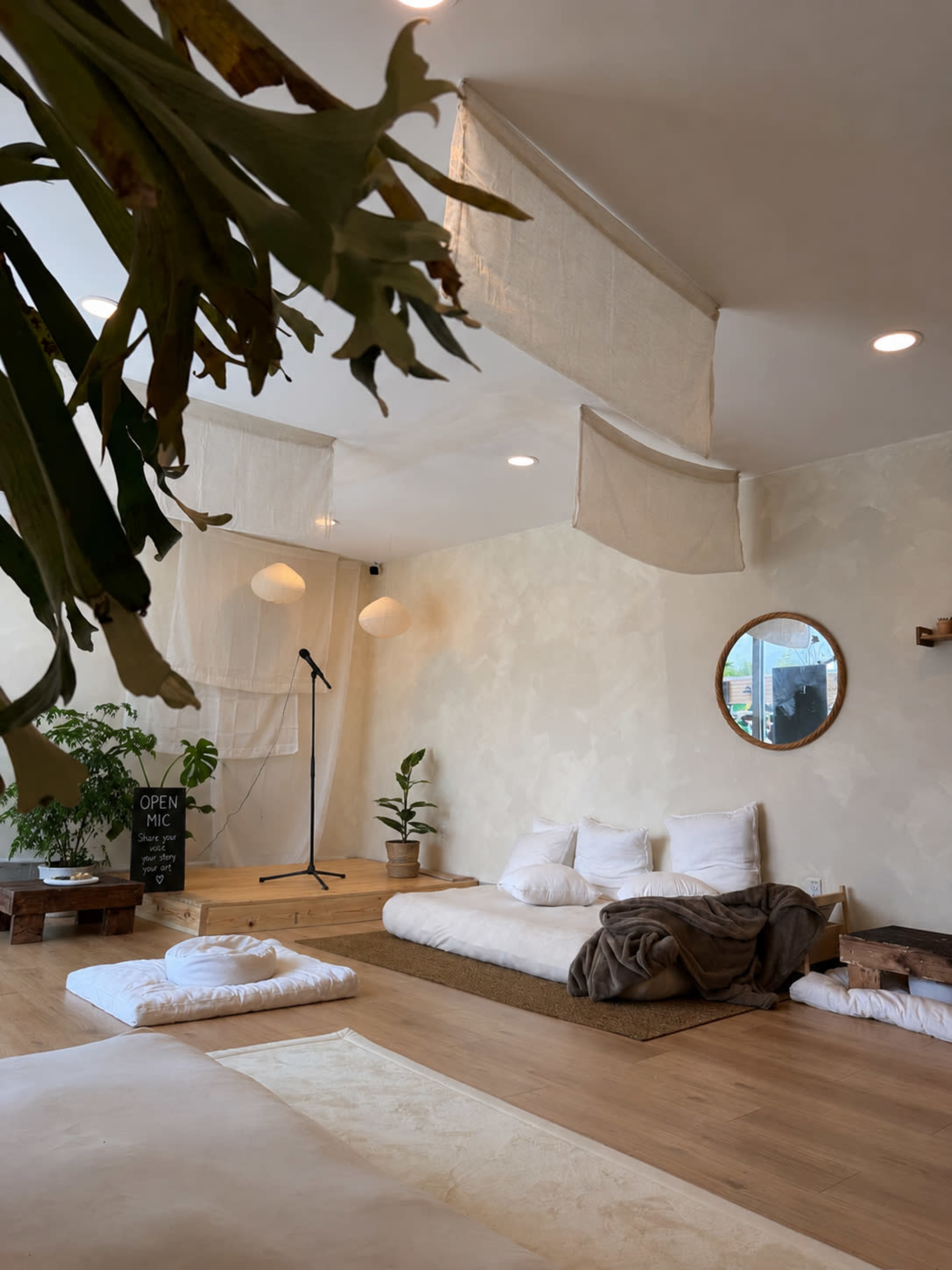 The image shows a minimalist, cozy interior space with cushions, plants, and a microphone set up for an open mic event.