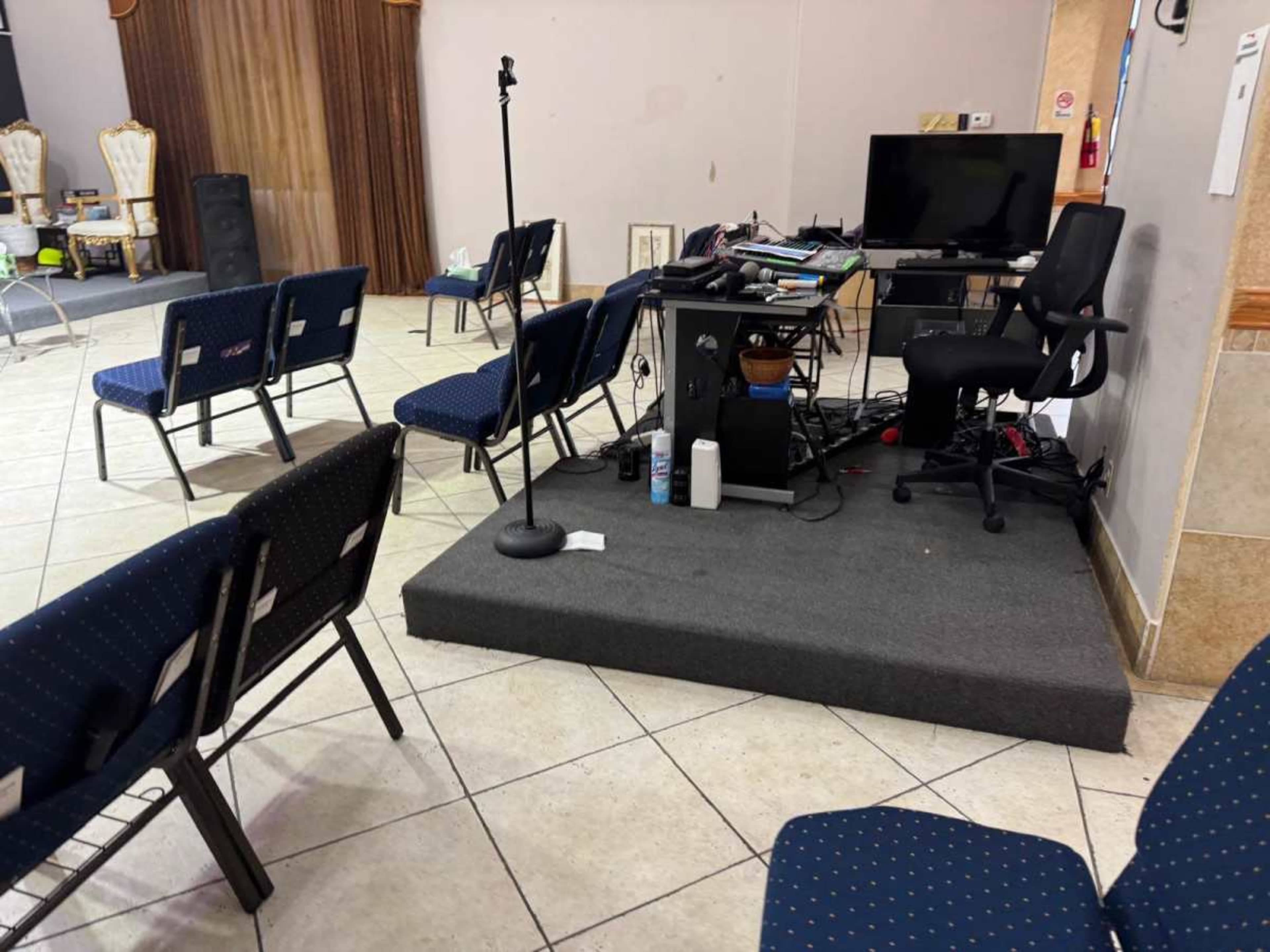 The image shows a small performance area with a microphone stand, a black desk cluttered with equipment, and blue chairs arranged in a semi-circle on tiled flooring.