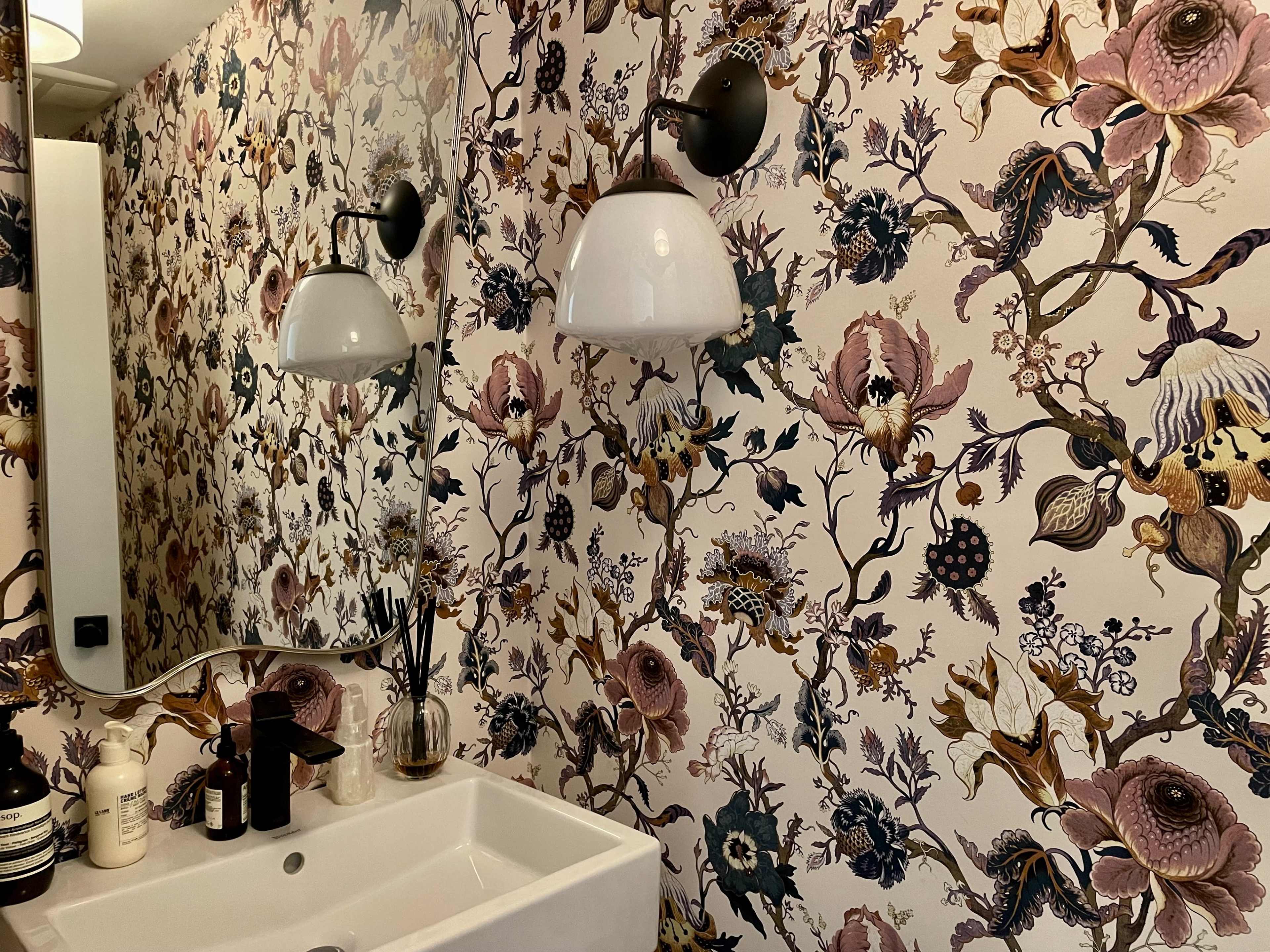The image shows a bathroom with floral-patterned wallpaper, a white sink, and wall-mounted light fixtures.