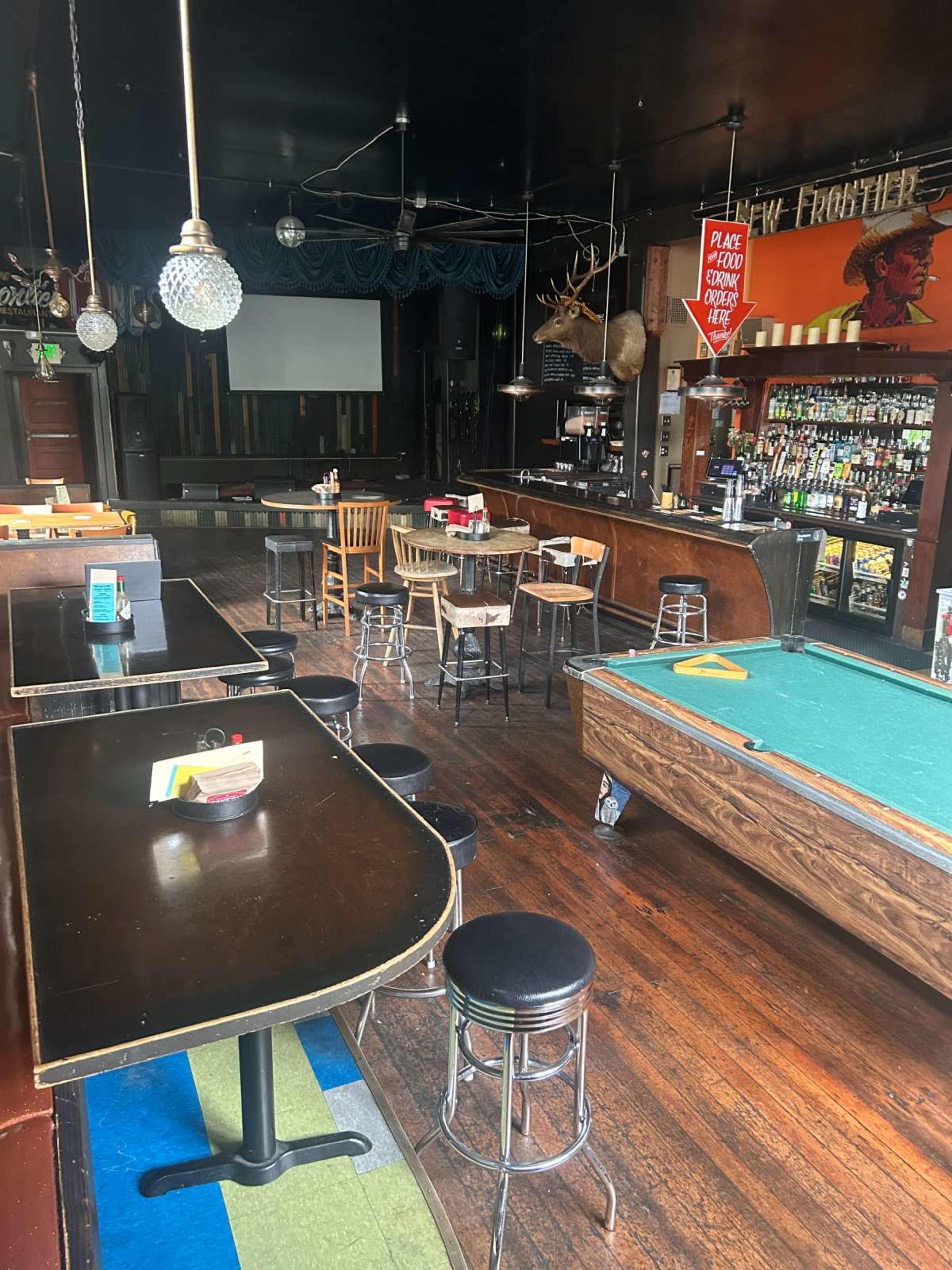 The image shows the interior of a bar with dark wood furnishings, multiple tables, a pool table, and a well-stocked bar counter.