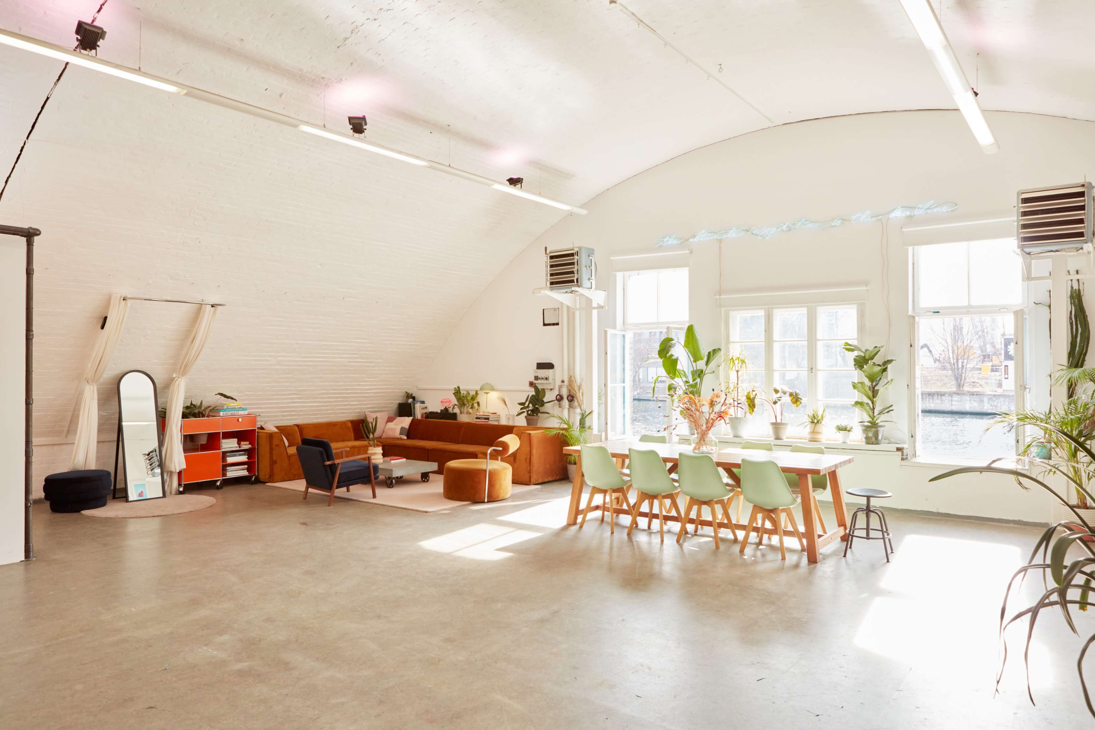 A spacious, well-lit room with a curved ceiling, featuring a seating area with a sofa, a dining table, and large windows filled with plants.