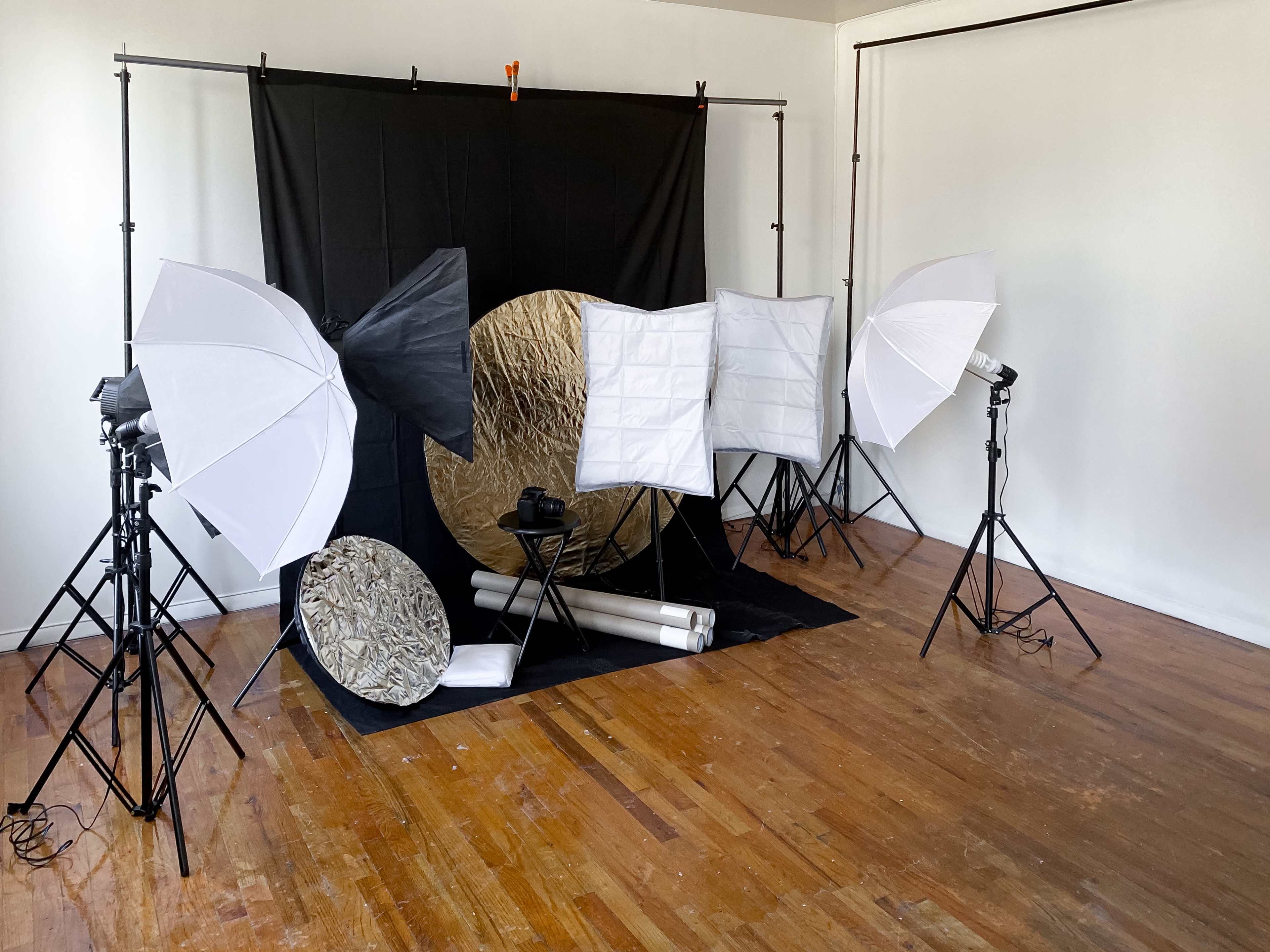A photo studio setup featuring a black backdrop, multiple light stands with softboxes, and reflector discs arranged on a wooden floor.