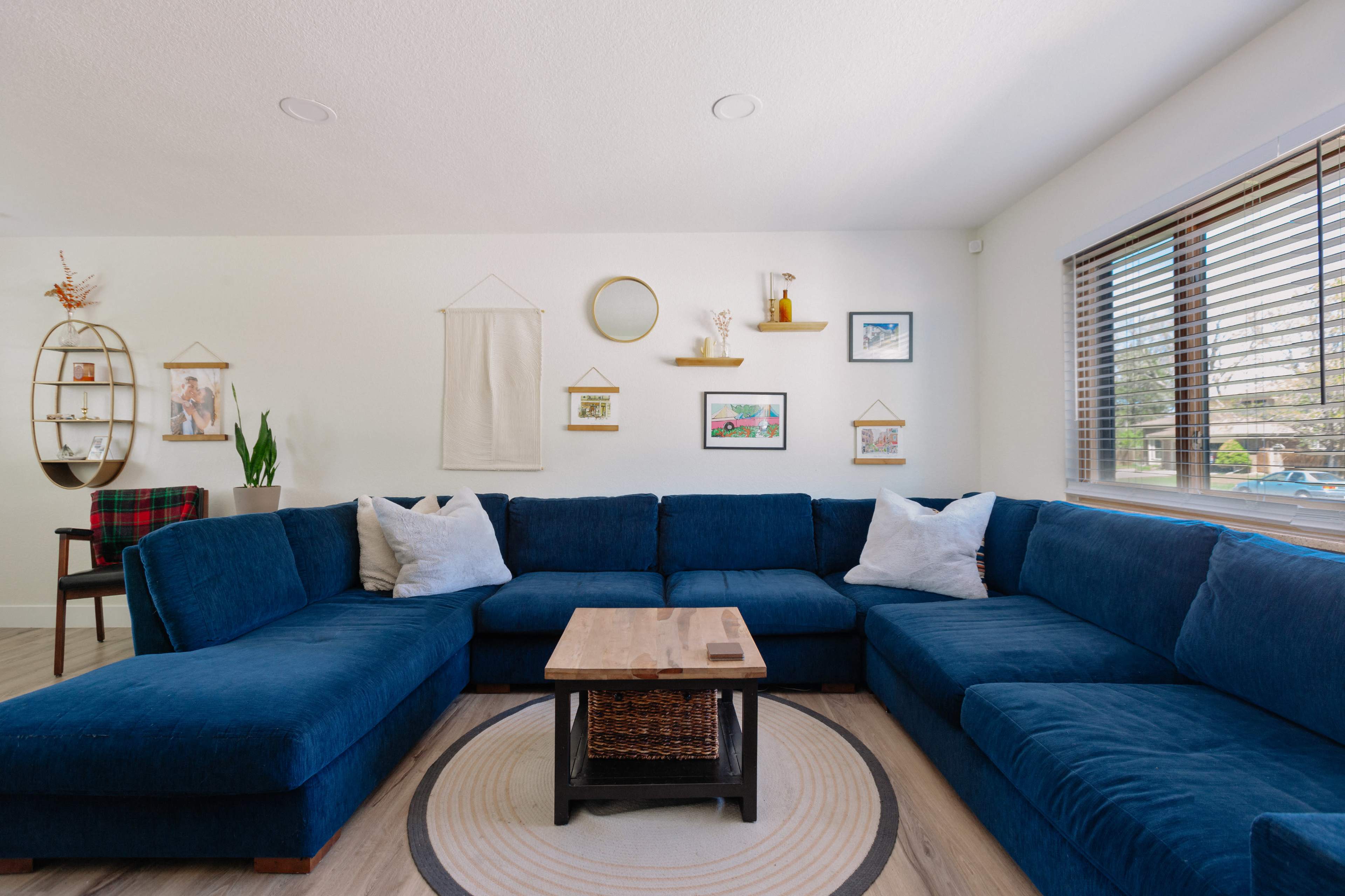 A spacious living room features a large blue sectional sofa, a wooden coffee table, and framed artwork on the walls beside a window with blinds.