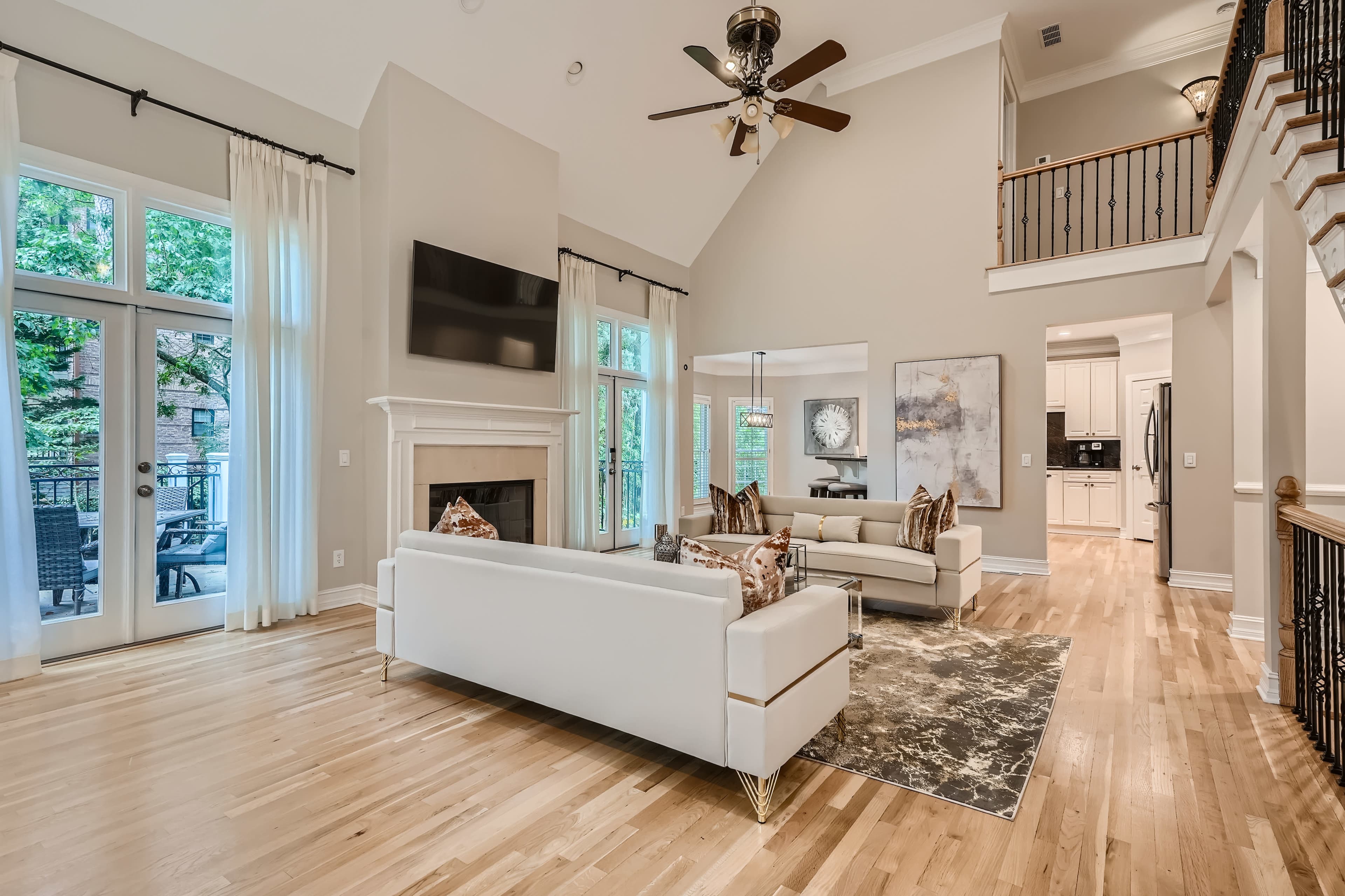 A spacious living room with white sofas, a large flat-screen TV mounted on the wall, and hardwood flooring, illuminated by natural light from large windows.