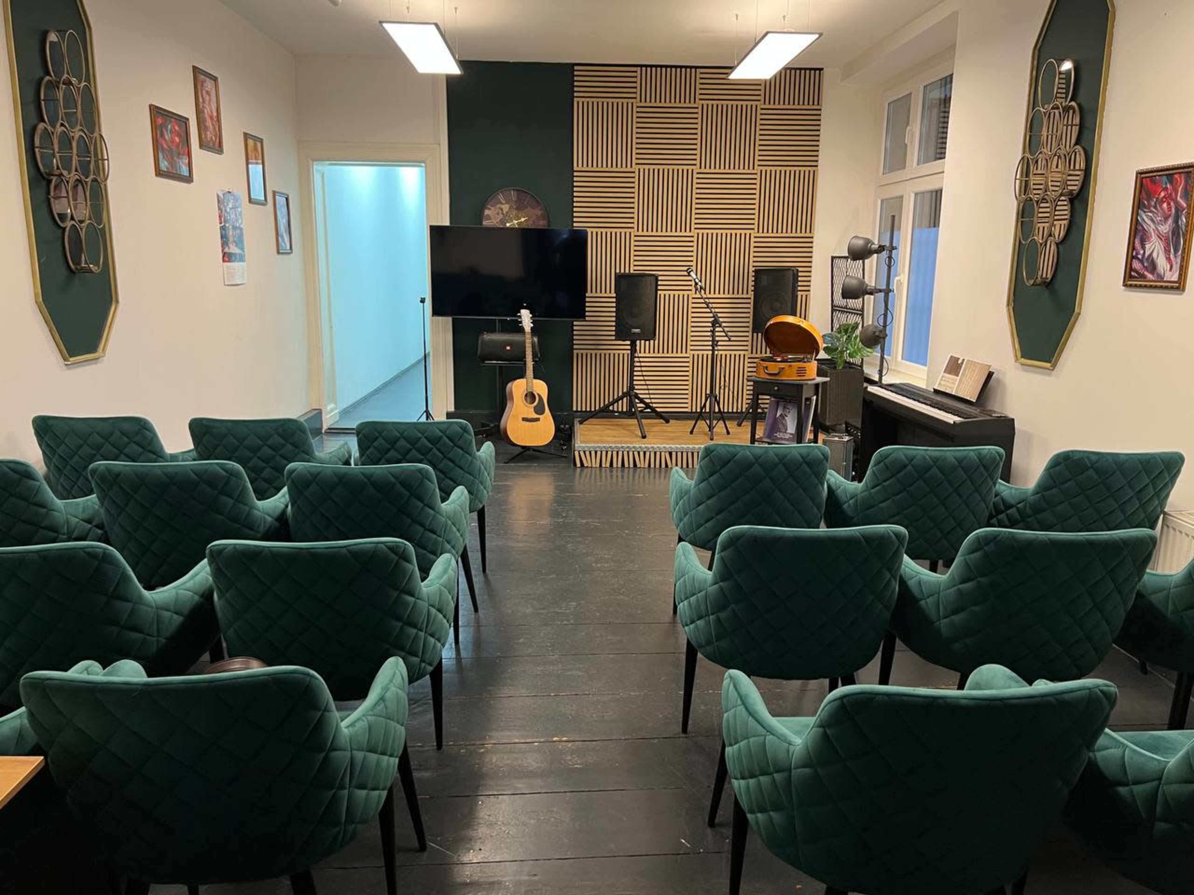 The image shows a performance space with green upholstered chairs arranged in rows facing a stage that features a guitar, a microphone stand, and a vintage-style record player.