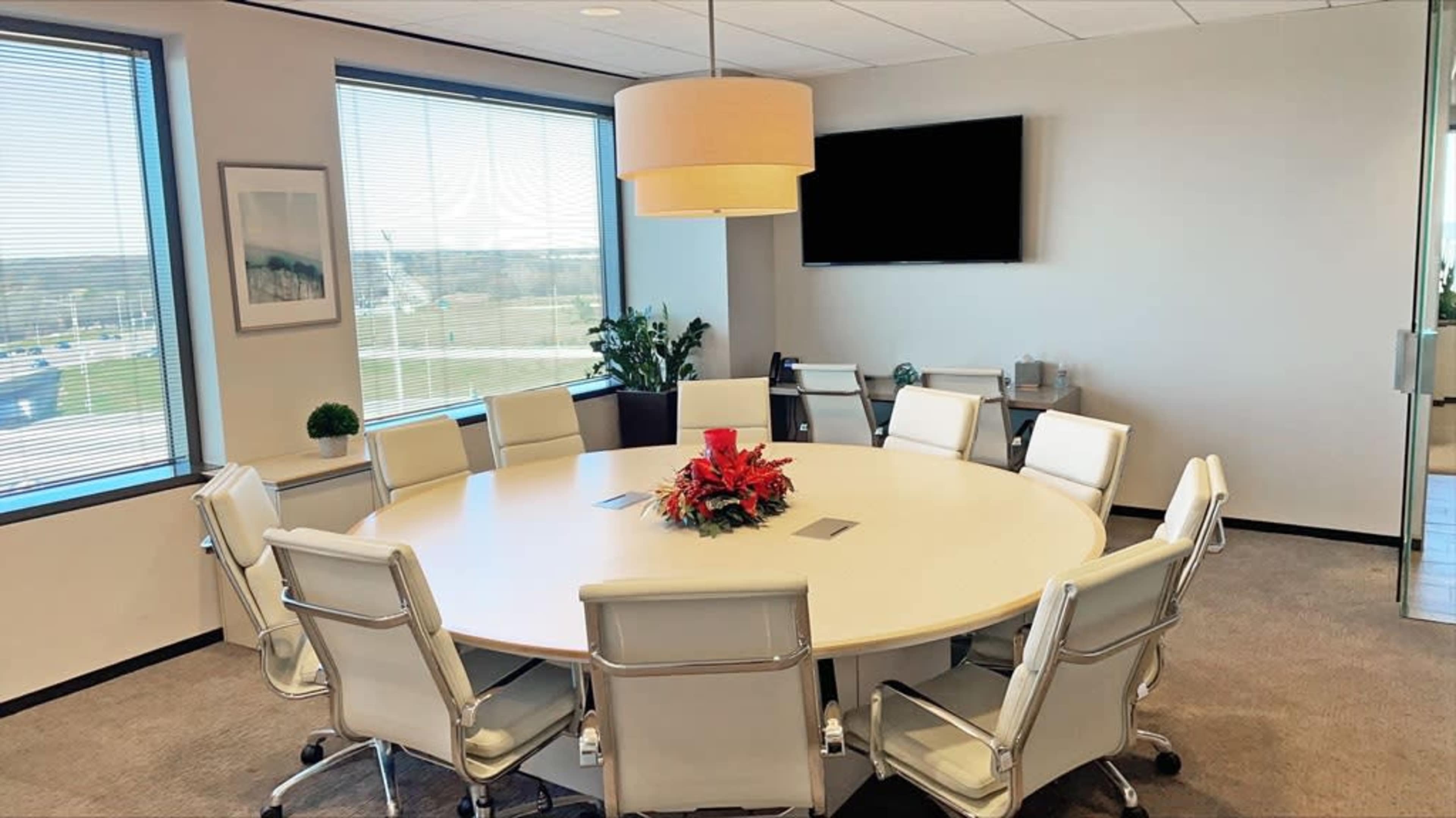 A round conference table surrounded by white chairs is set up in a well-lit meeting room with large windows displaying an outdoor view.