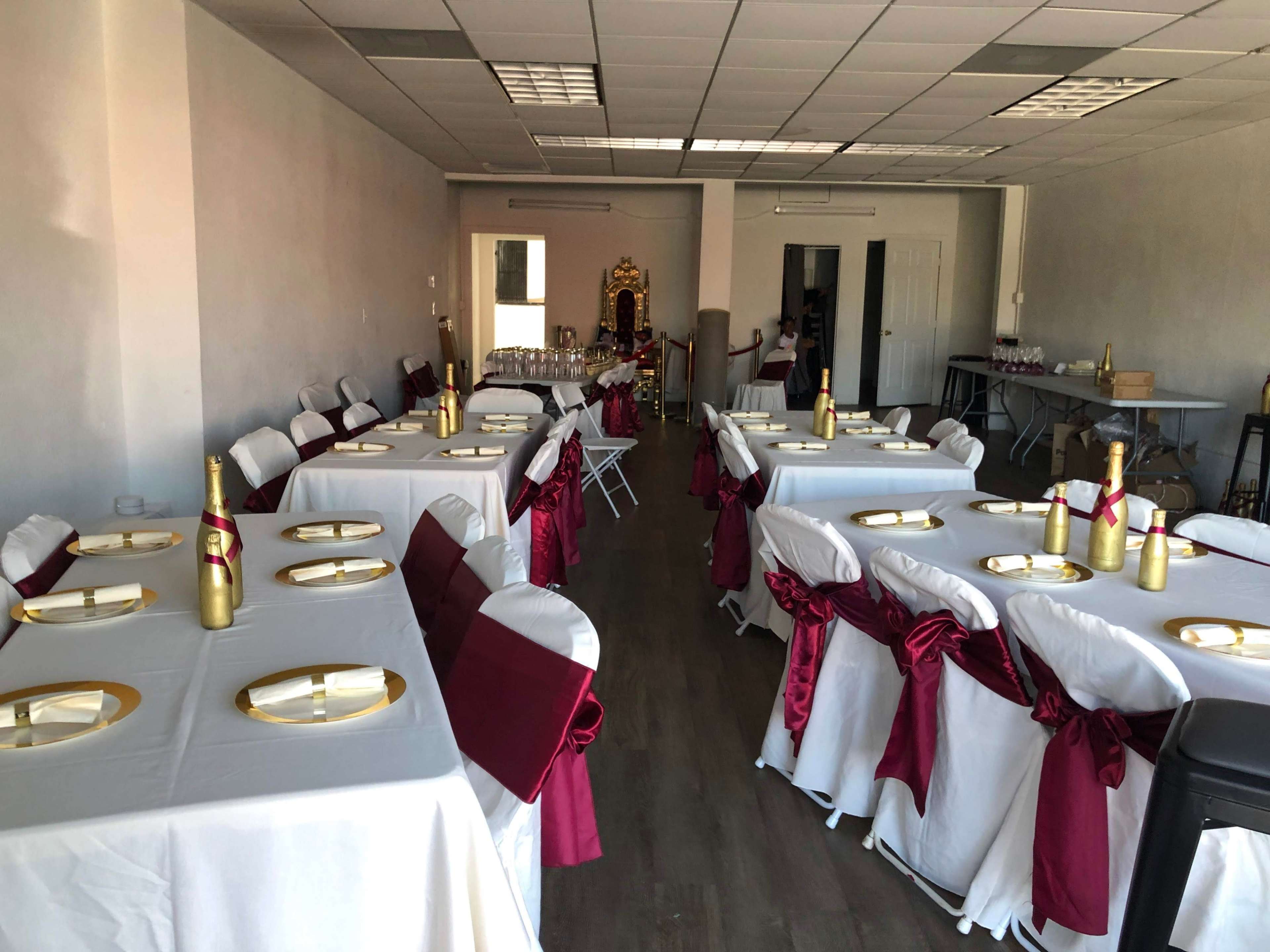 A banquet hall with several tables arranged in a row, each covered with white tablecloths and adorned with golden plates and maroon ribbons on the chairs.