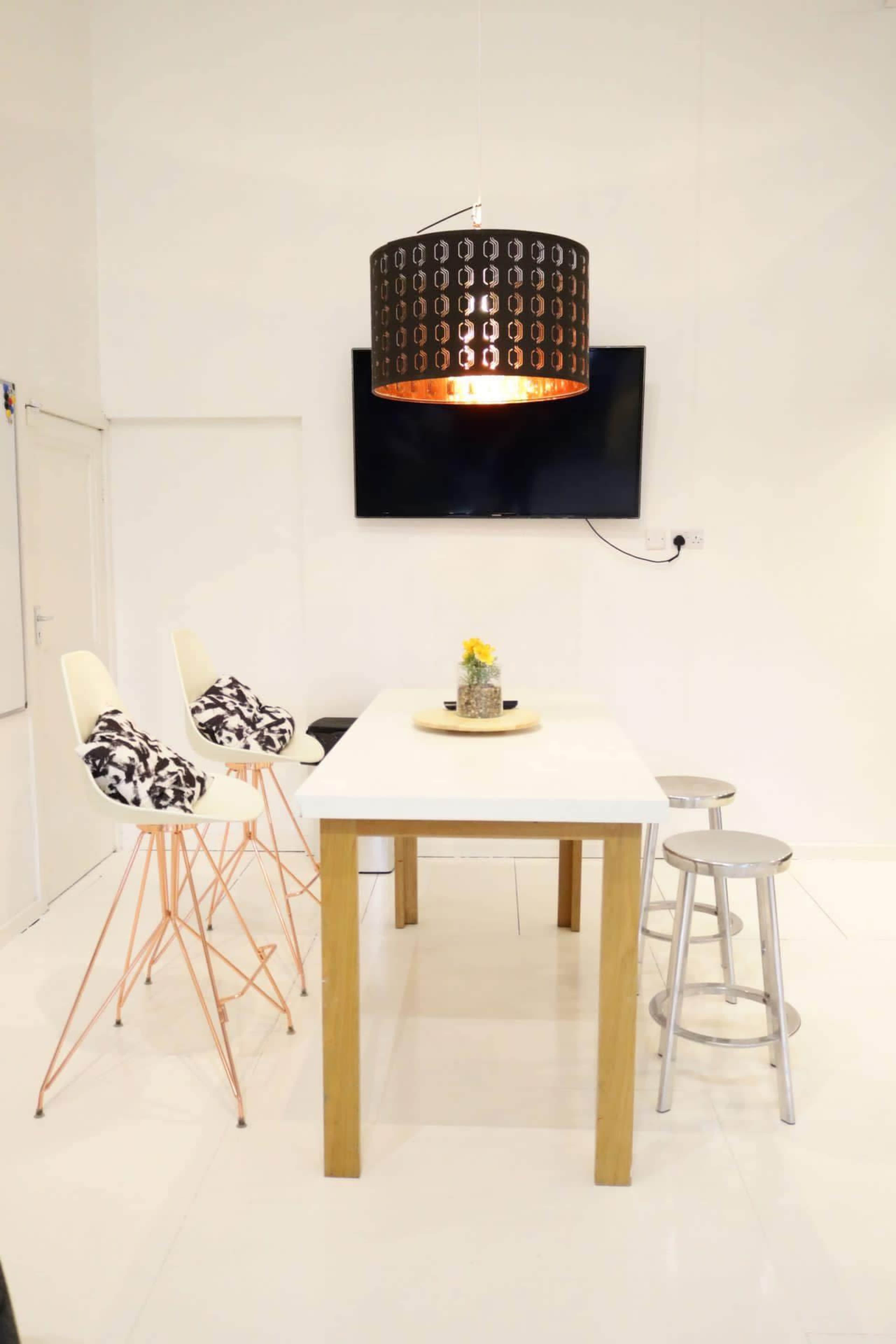 The image shows a modern dining area with a white table, bar stools, a decorative pendant light, and a wall-mounted television.