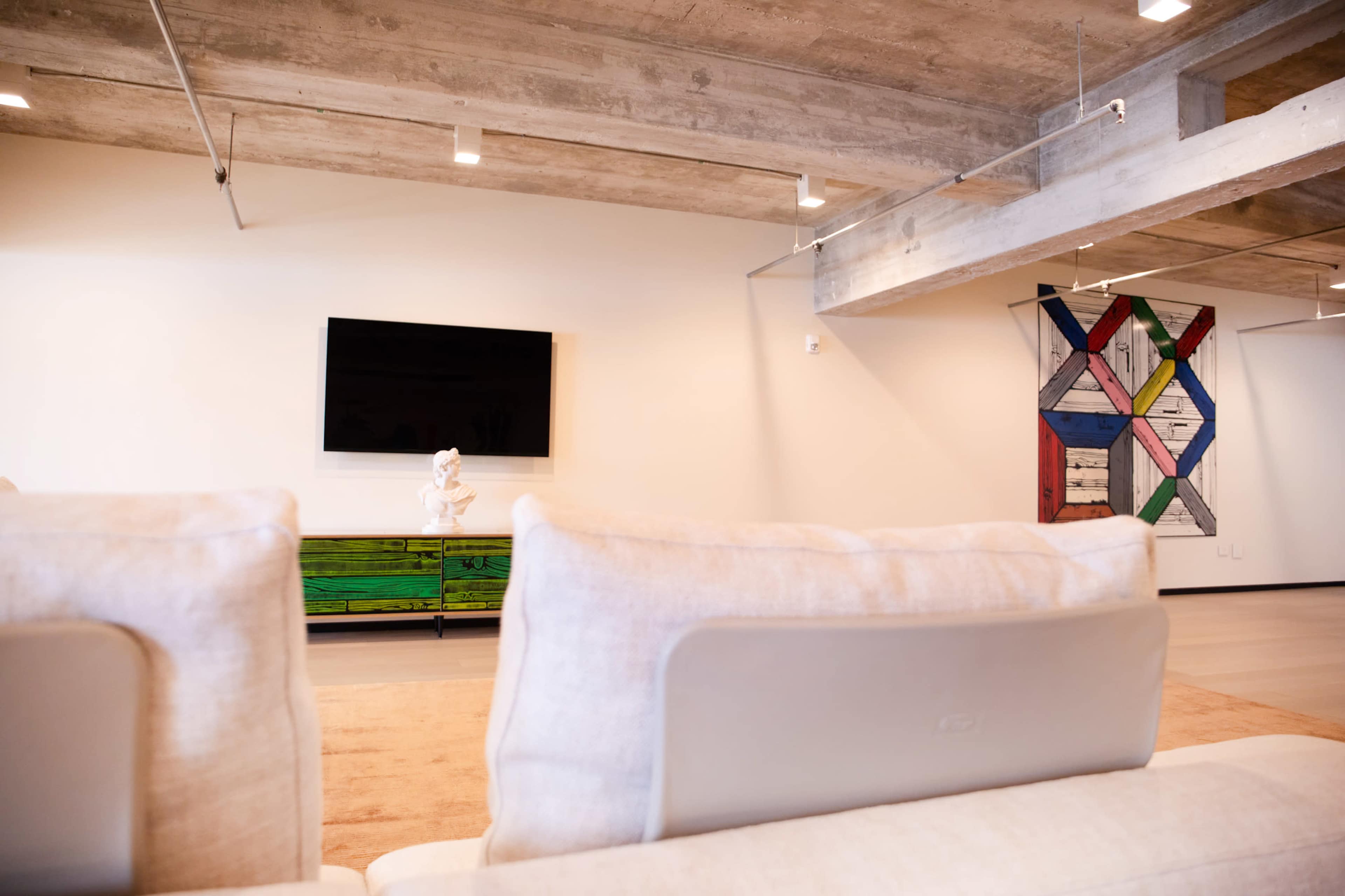 The image shows a modern living room with a flat-screen TV mounted on the wall, a green wooden media console, and an abstract wall art piece.