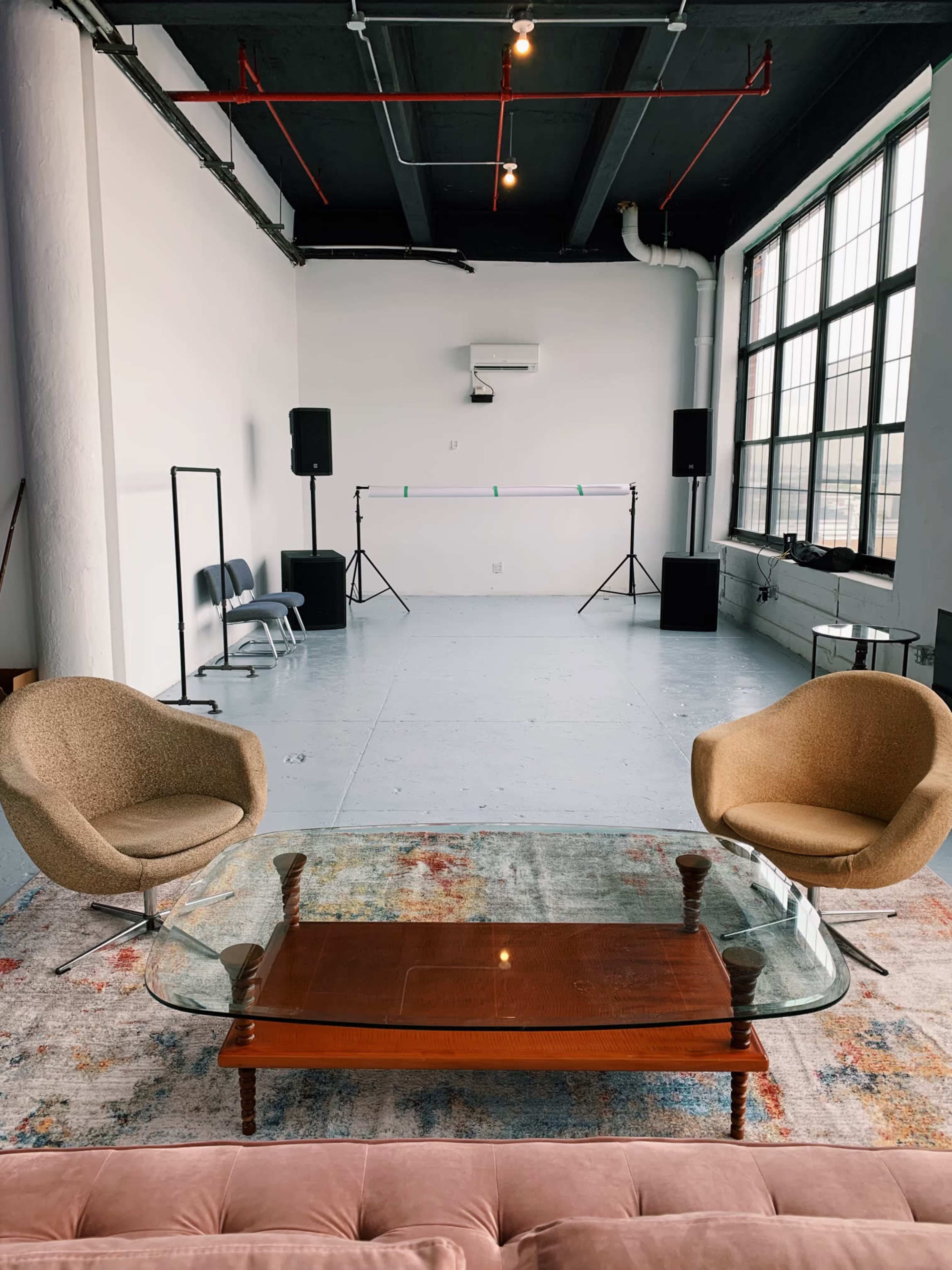 A spacious room features a glass coffee table, two armchairs, and a stage setup with speakers and a backdrop.