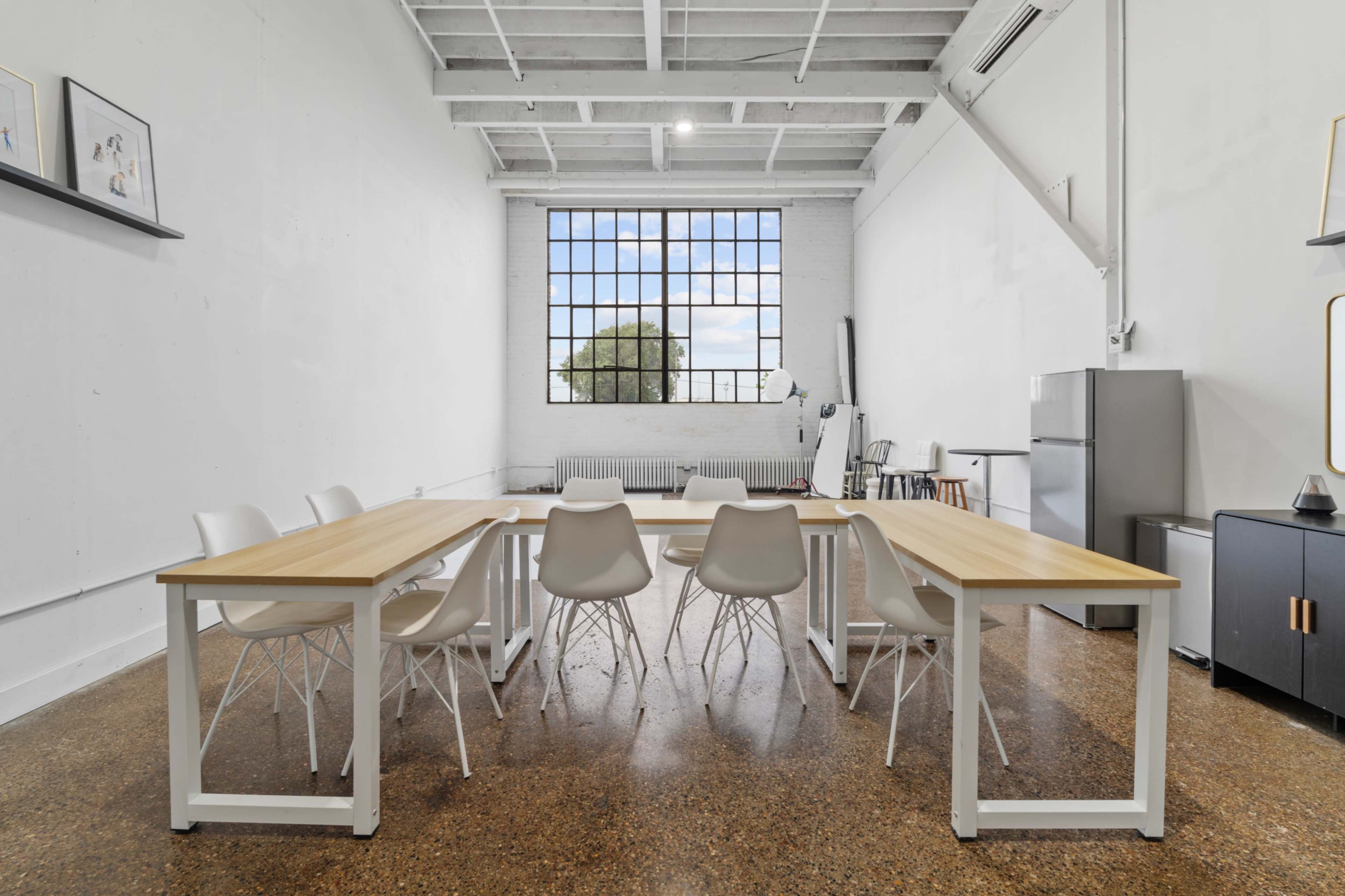 A long wooden table with white chairs is set up in a spacious room featuring a large window and minimal furnishings.