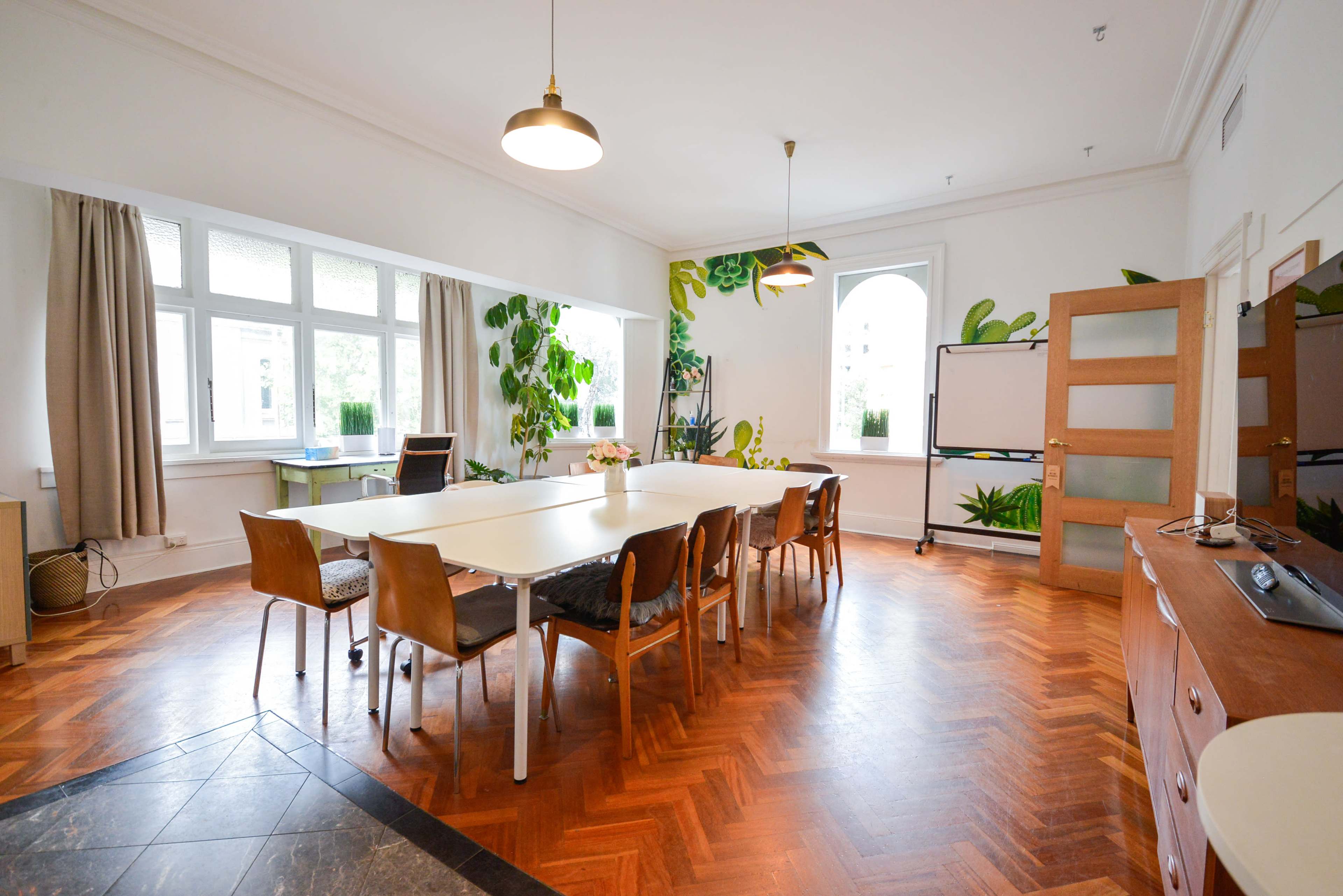 A spacious meeting room features a large table surrounded by chairs, with large windows and plants decorating the walls.