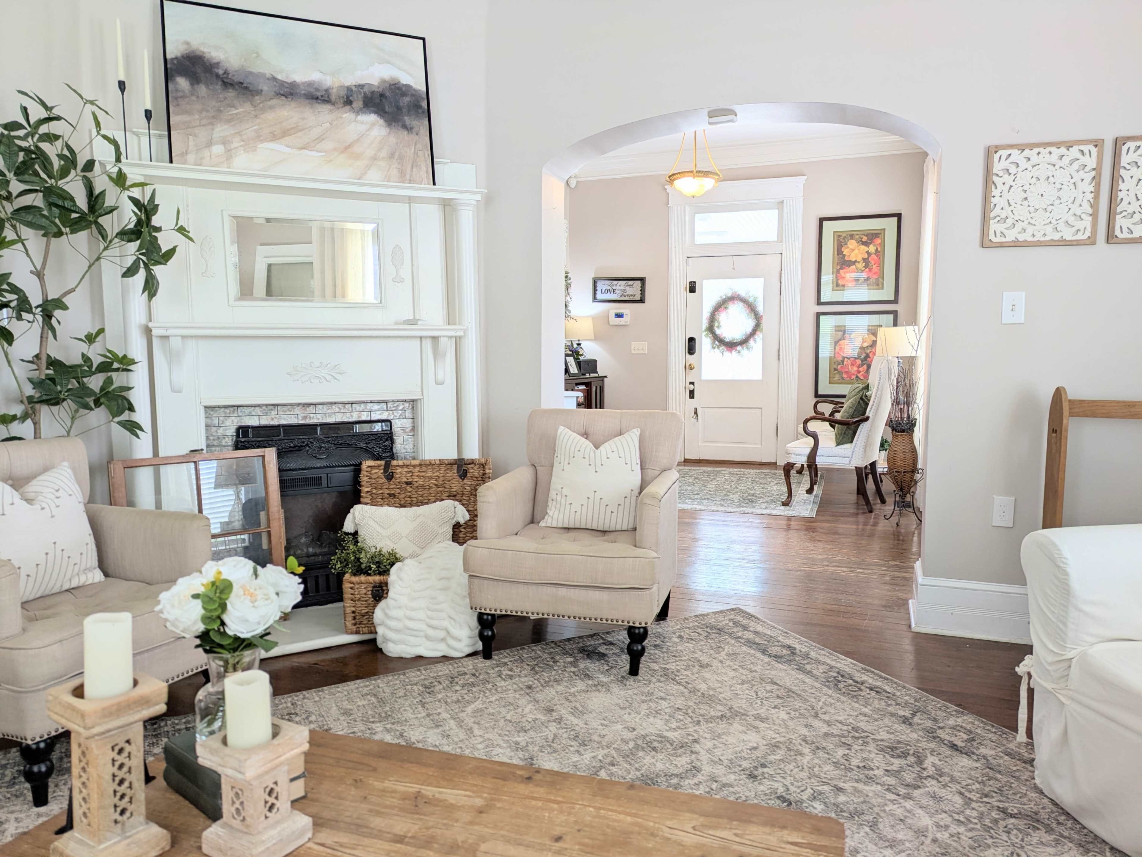 The image shows a cozy living room with two upholstered chairs, a coffee table, and a fireplace, leading to an entrance with a wreath on the door.