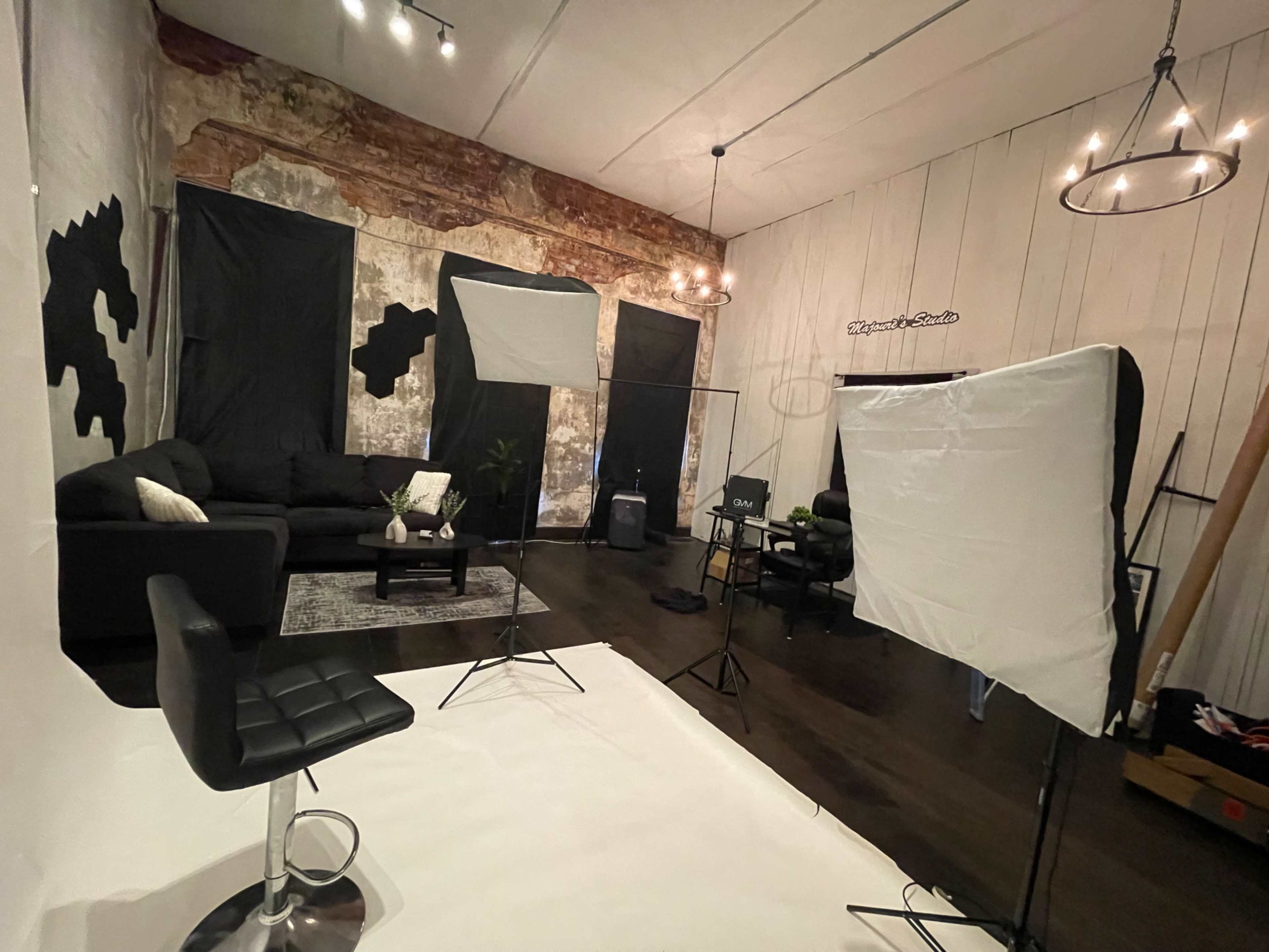 The image shows a photography studio with a sectional couch, black fabric backdrops, and lighting equipment set up for a shoot.