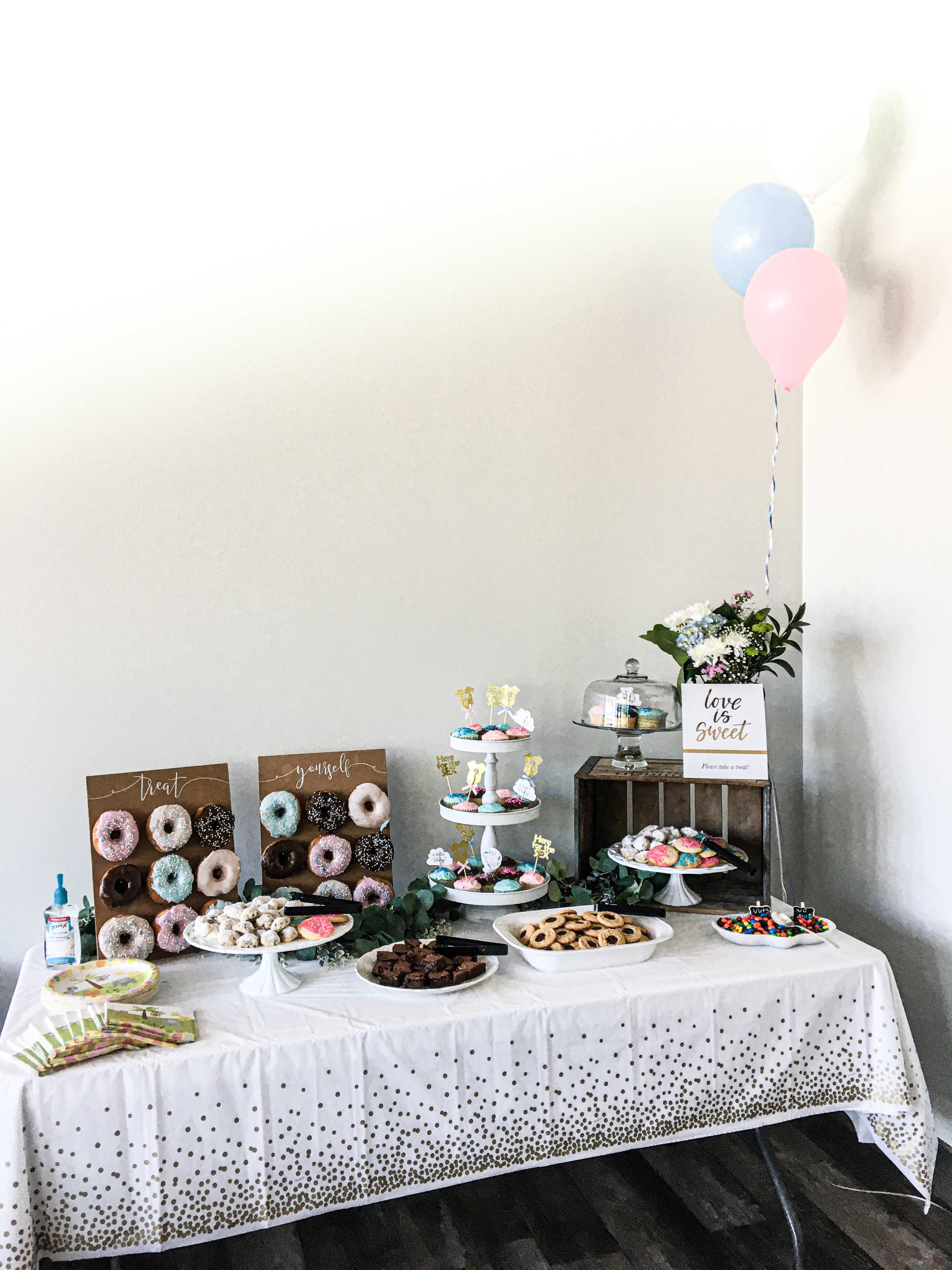 A decorated table is set up with an array of desserts, including donuts, cupcakes, cookies, and snacks, alongside colorful balloons and floral arrangements.