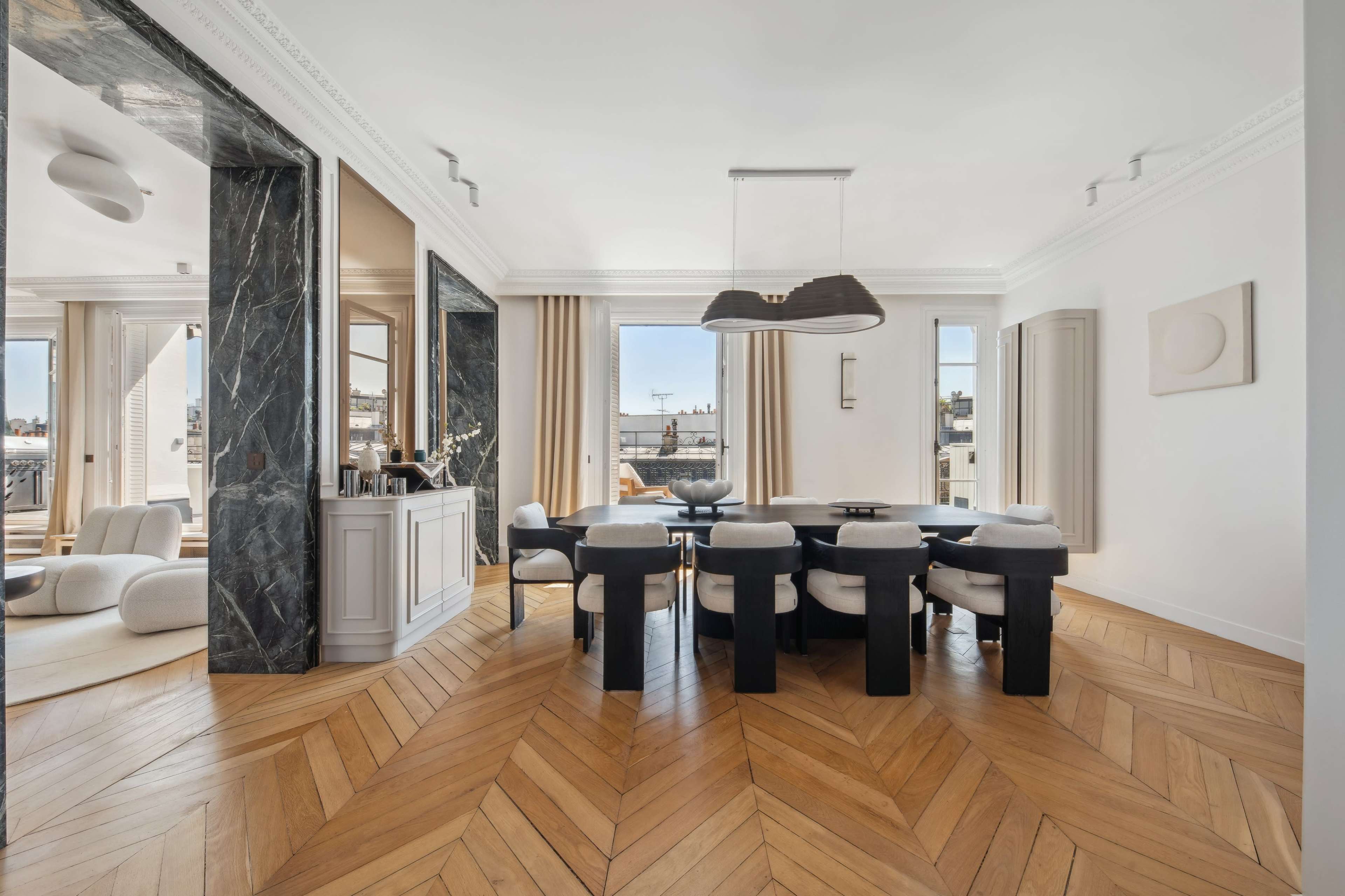 A modern dining room with a large black table surrounded by upholstered chairs, hardwood herringbone flooring, and large windows offering a view of the city skyline.
