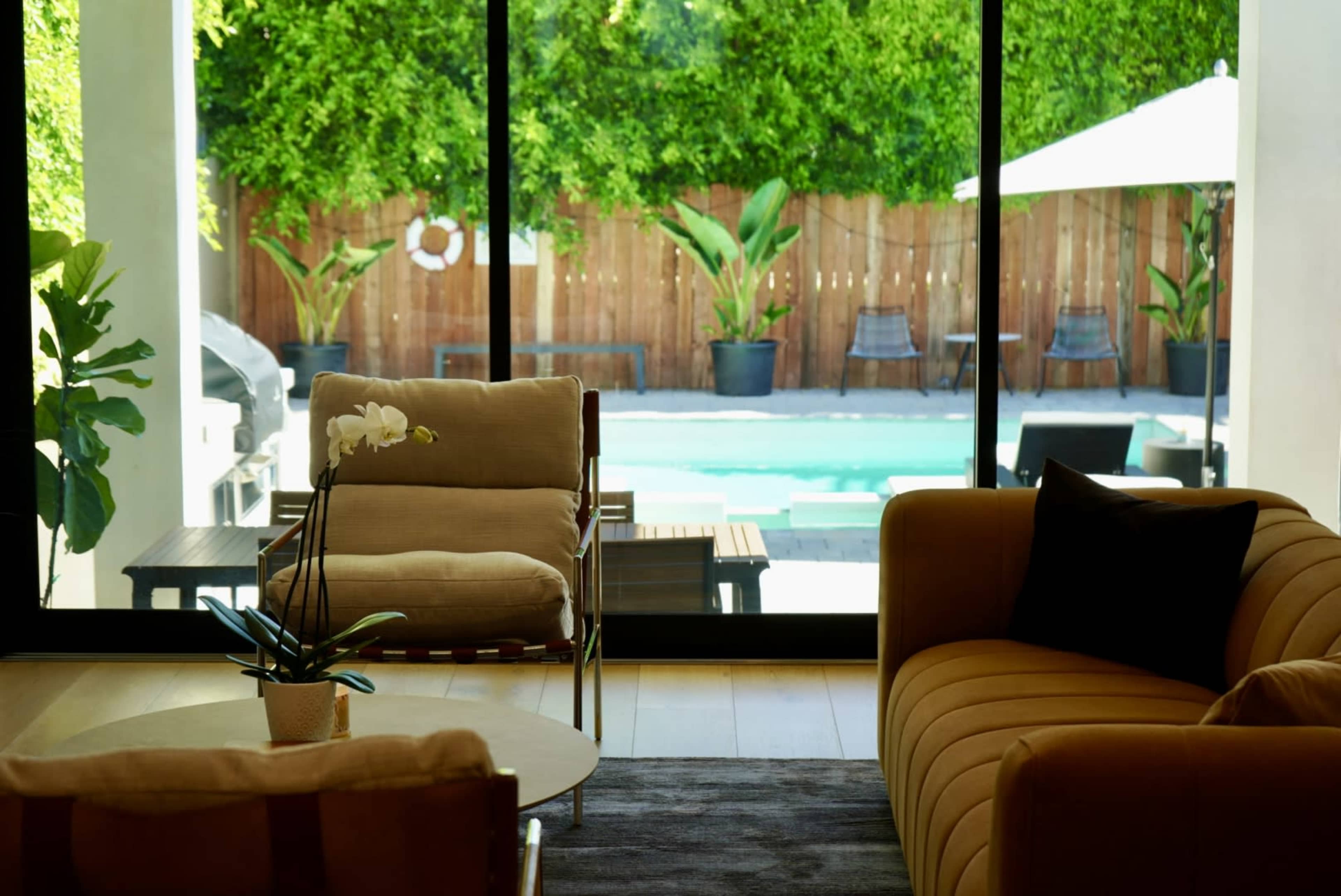 The image shows a modern living room with a view of a pool area surrounded by greenery through large glass doors.