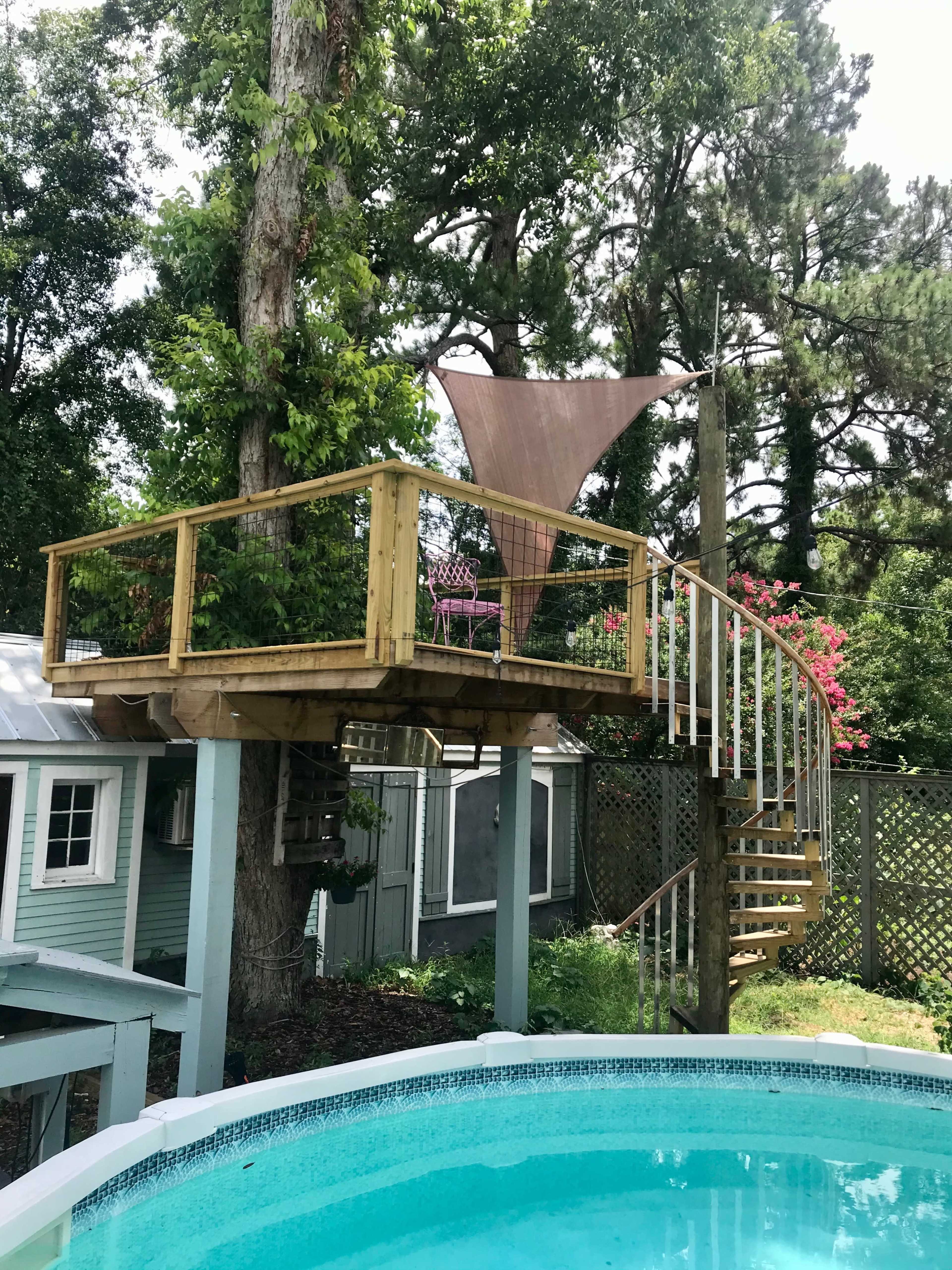 A wooden platform with a spiral staircase is positioned above a swimming pool, surrounded by trees and colorful flowers.