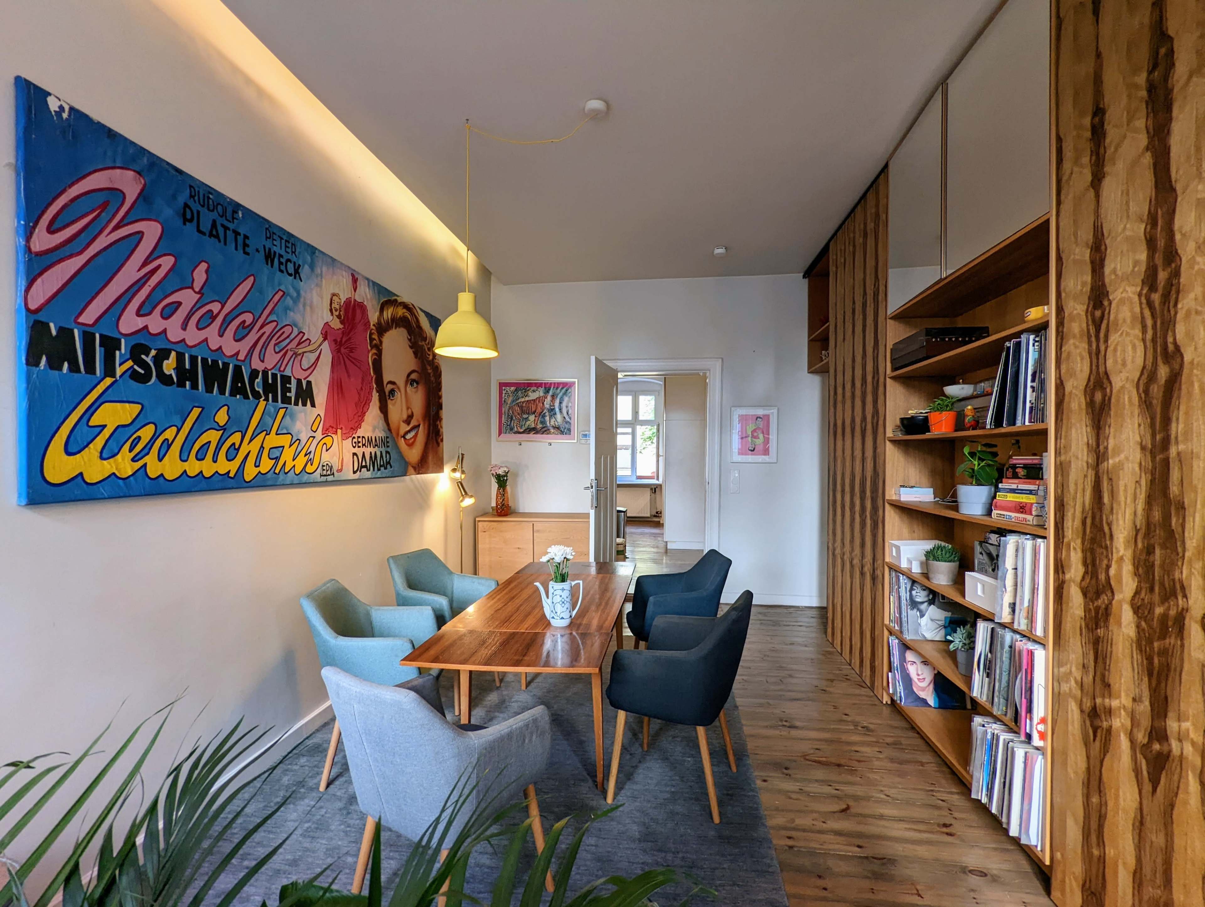 A modern dining area featuring a long wooden table surrounded by chairs, colorful wall art, and a bookshelf filled with books.