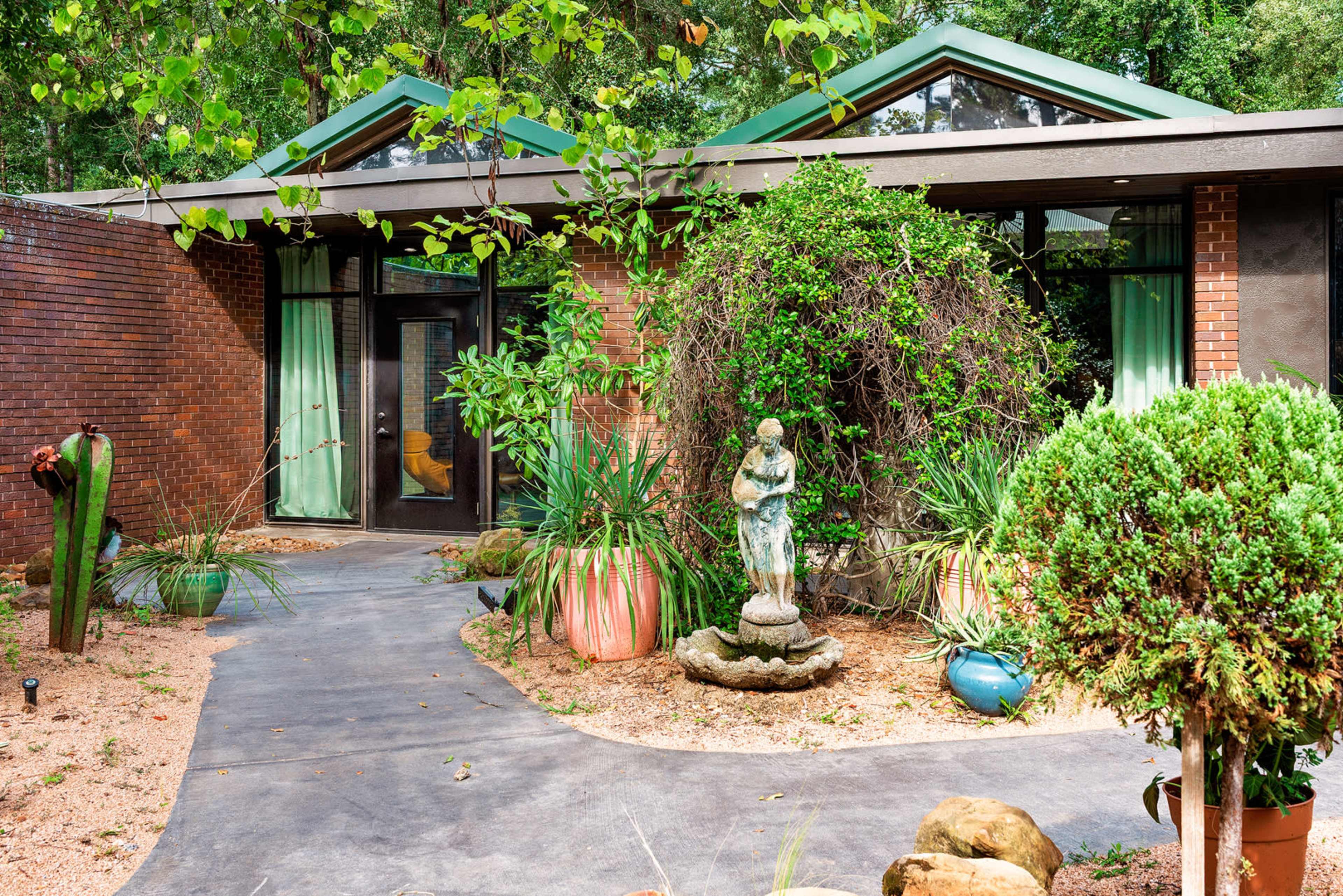The image shows a modern house with a landscaped front yard featuring a pathway, plants, and a small statue in a fountain.