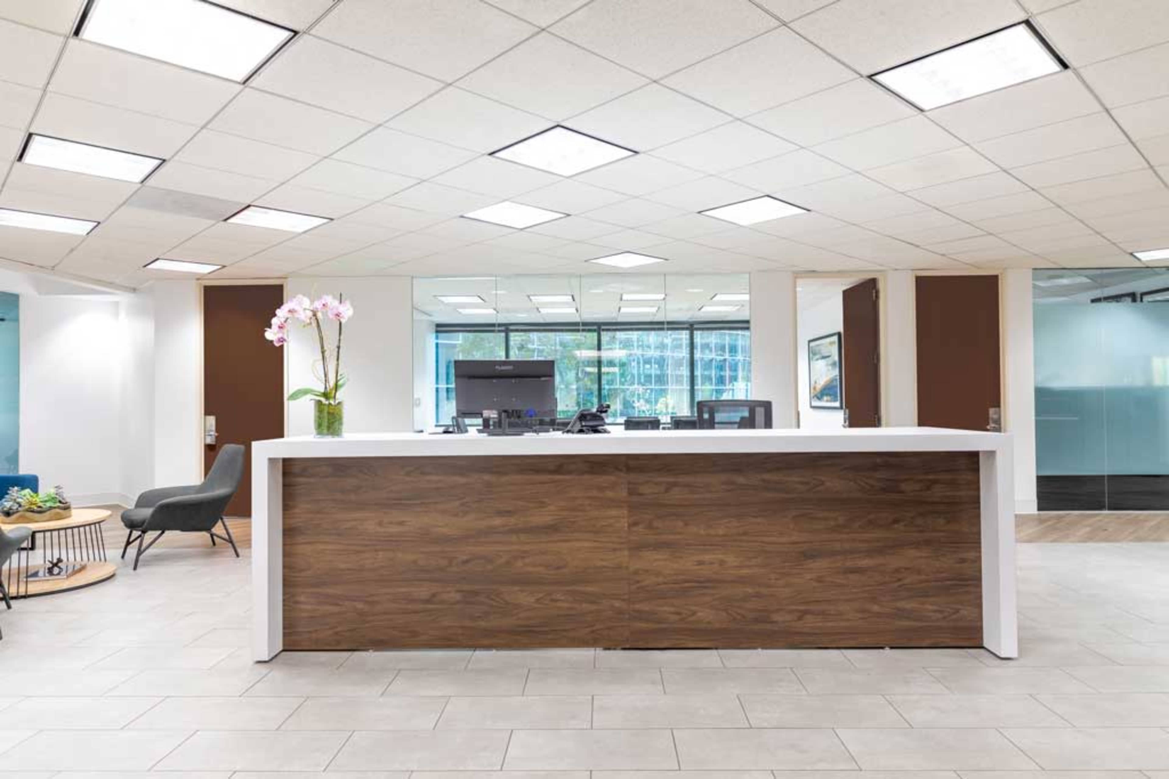 The image shows a modern reception area with a wooden front desk, a plant, and office space visible in the background.