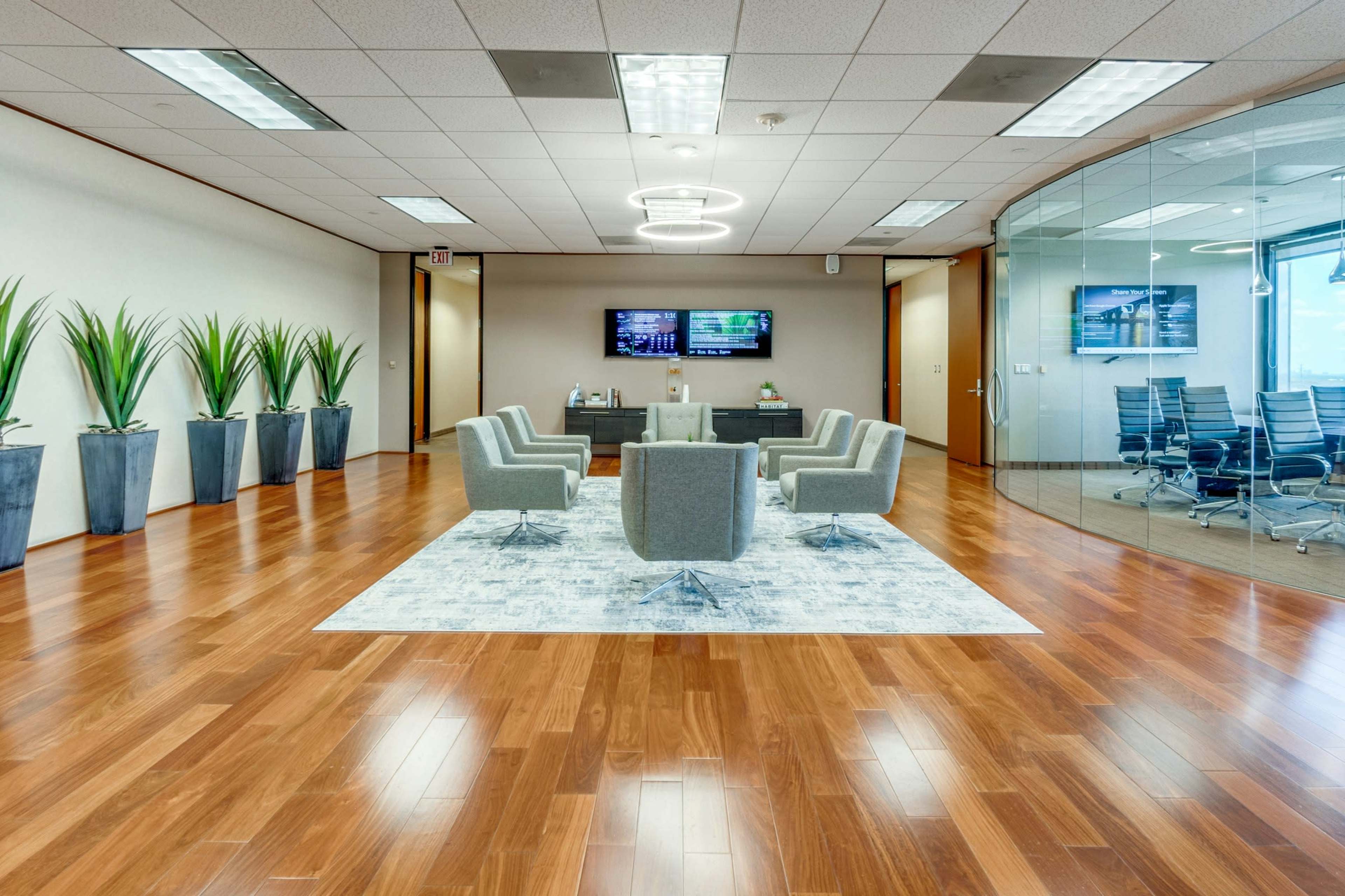 The image shows a modern office space featuring a seating area with four gray chairs arranged around a rug, surrounded by potted plants and a glass-walled conference room.
