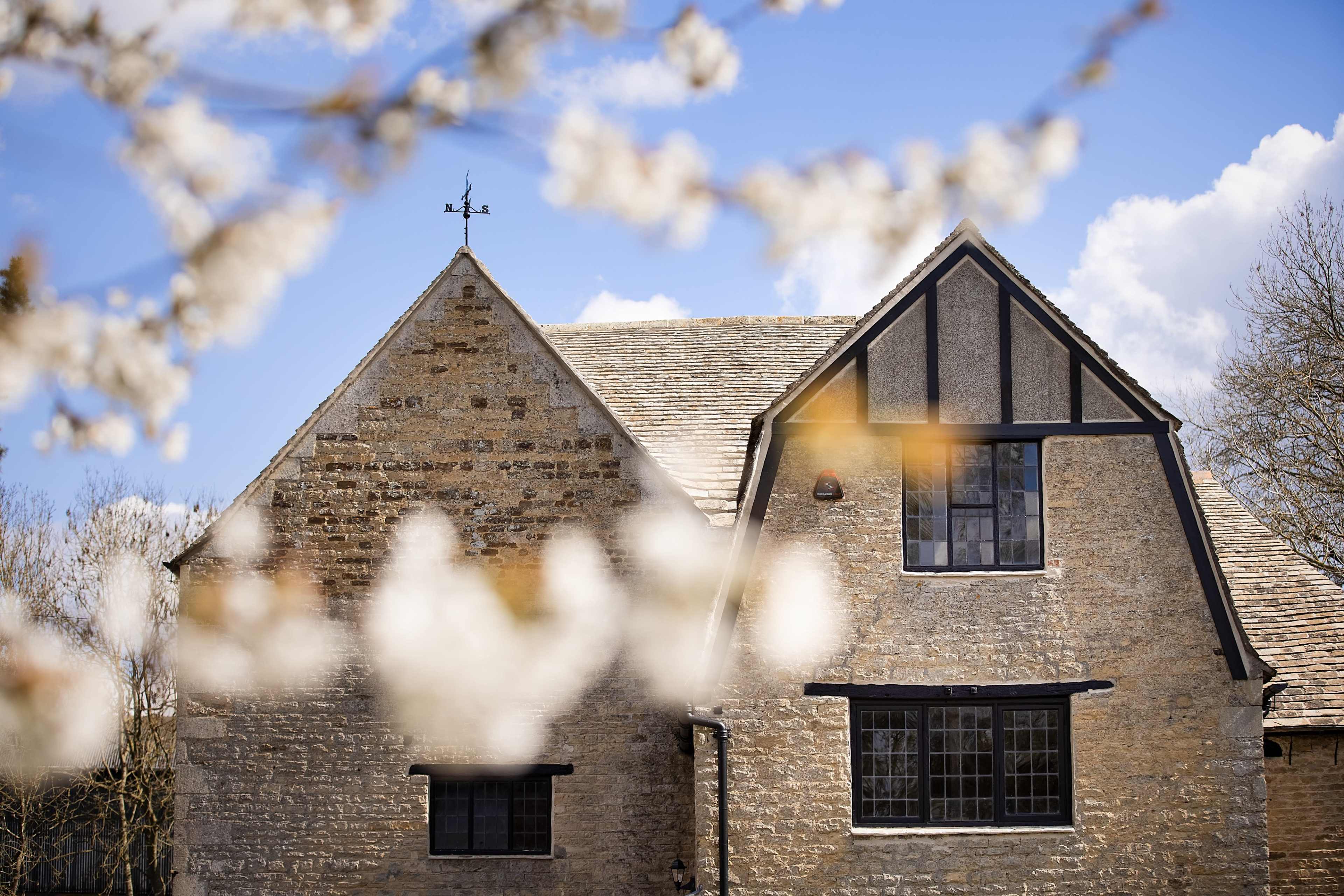 A stone house with a gable roof and dark window frames stands against a blue sky, framed by flowering branches in the foreground.